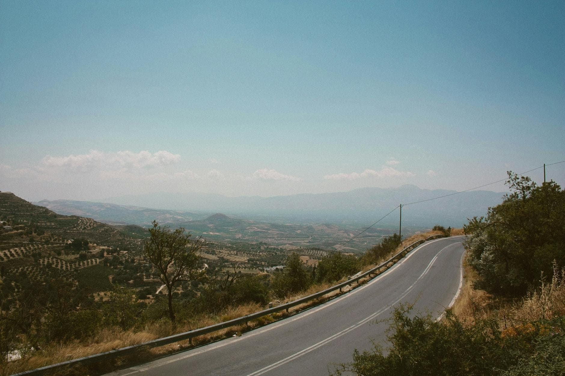 Kurvenreiche Straße mit Leitplanken durch eine offene hügelige Landschaft mit Blick auf Täler und ferne Berge unter einem klaren blauen Himmel auf Kreta.