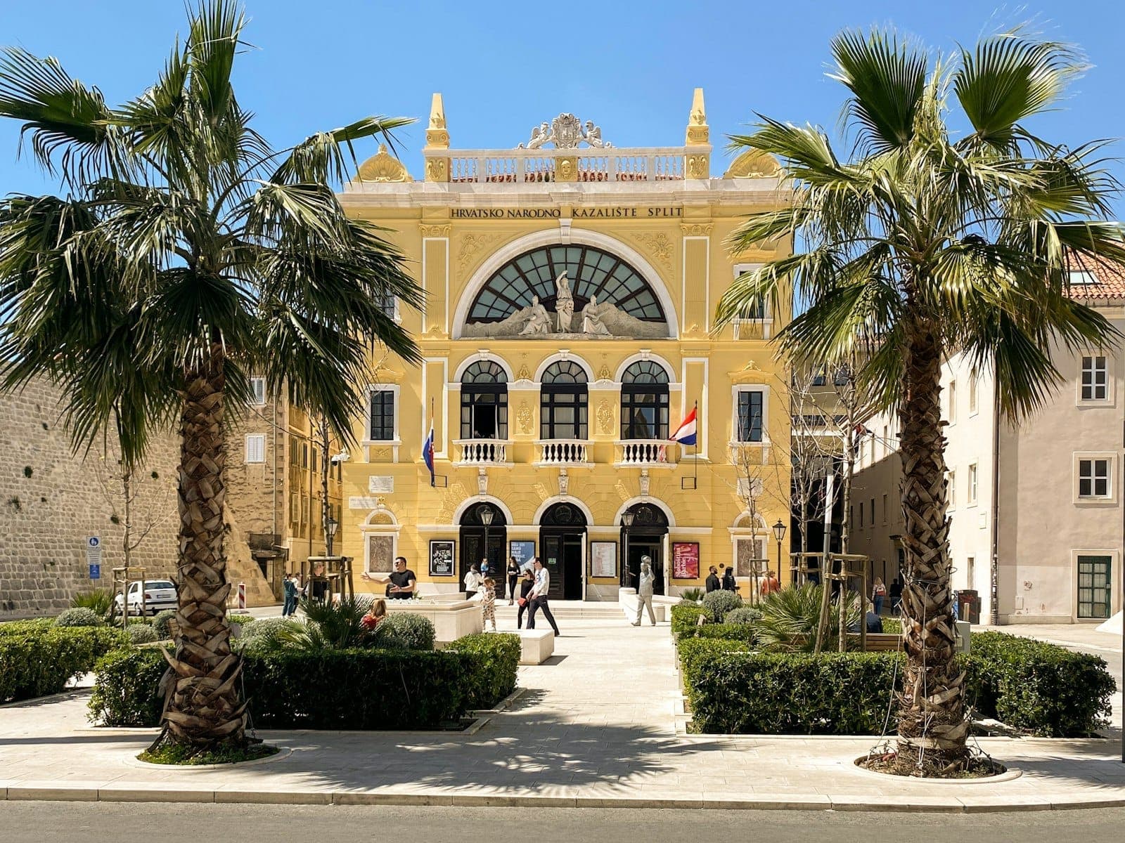 Facciata principale del Teatro Nazionale Croato di Spalato con esterno giallo, palme e persone che passeggiano sulla piazza in una giornata di sole.
