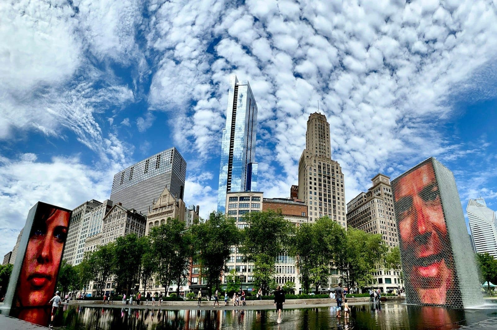 Crown Fountain in Chicago’s Millennium Park with digital faces on glass towers, reflecting pool, city skyscrapers, and people enjoying a sunny day.