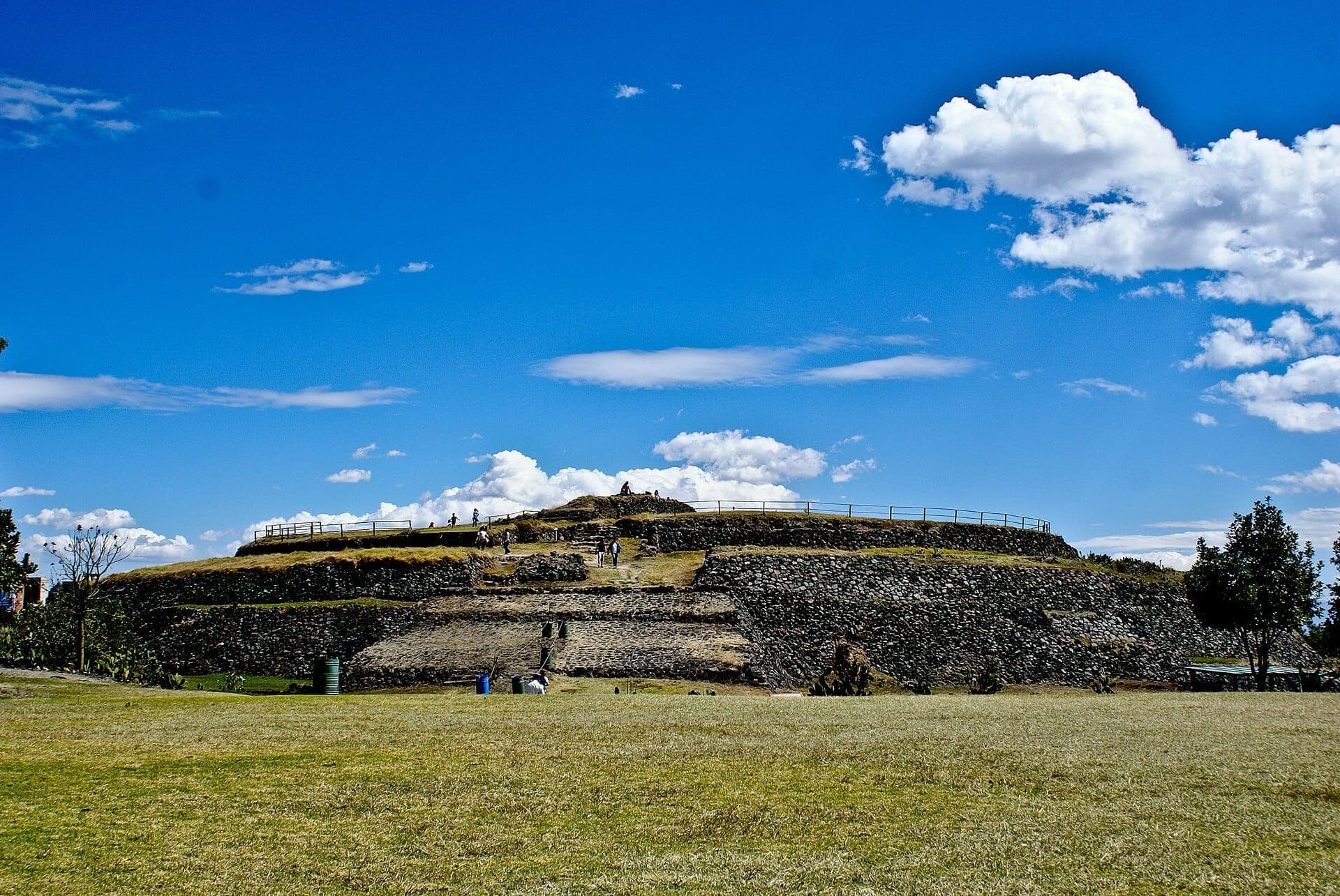 ภาพมุมกว้างของพีระมิดทรงกลมในเขตโบราณคดี Cuicuilco พร้อมลานหินขั้นบันได พื้นที่สนามหญ้า และนักท่องเที่ยวใต้ท้องฟ้าสีฟ้าสดใส