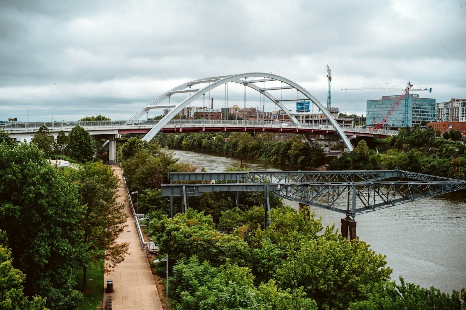 View of the pedestrian path at Cumberland Park in Nashville, with lush greenery, the river, and a modern arched bridge under clouded skies.