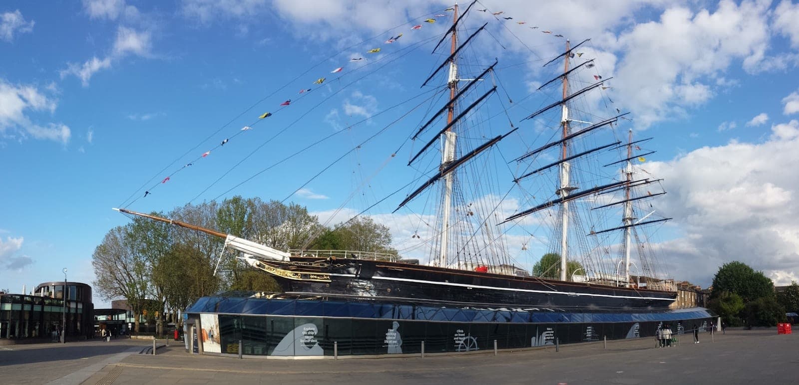 Der historische Teeklipper Cutty Sark im Hafen von Greenwich, umgeben von einer Glasplaza, unter einem strahlend blauen Himmel mit vereinzelten Wolken.