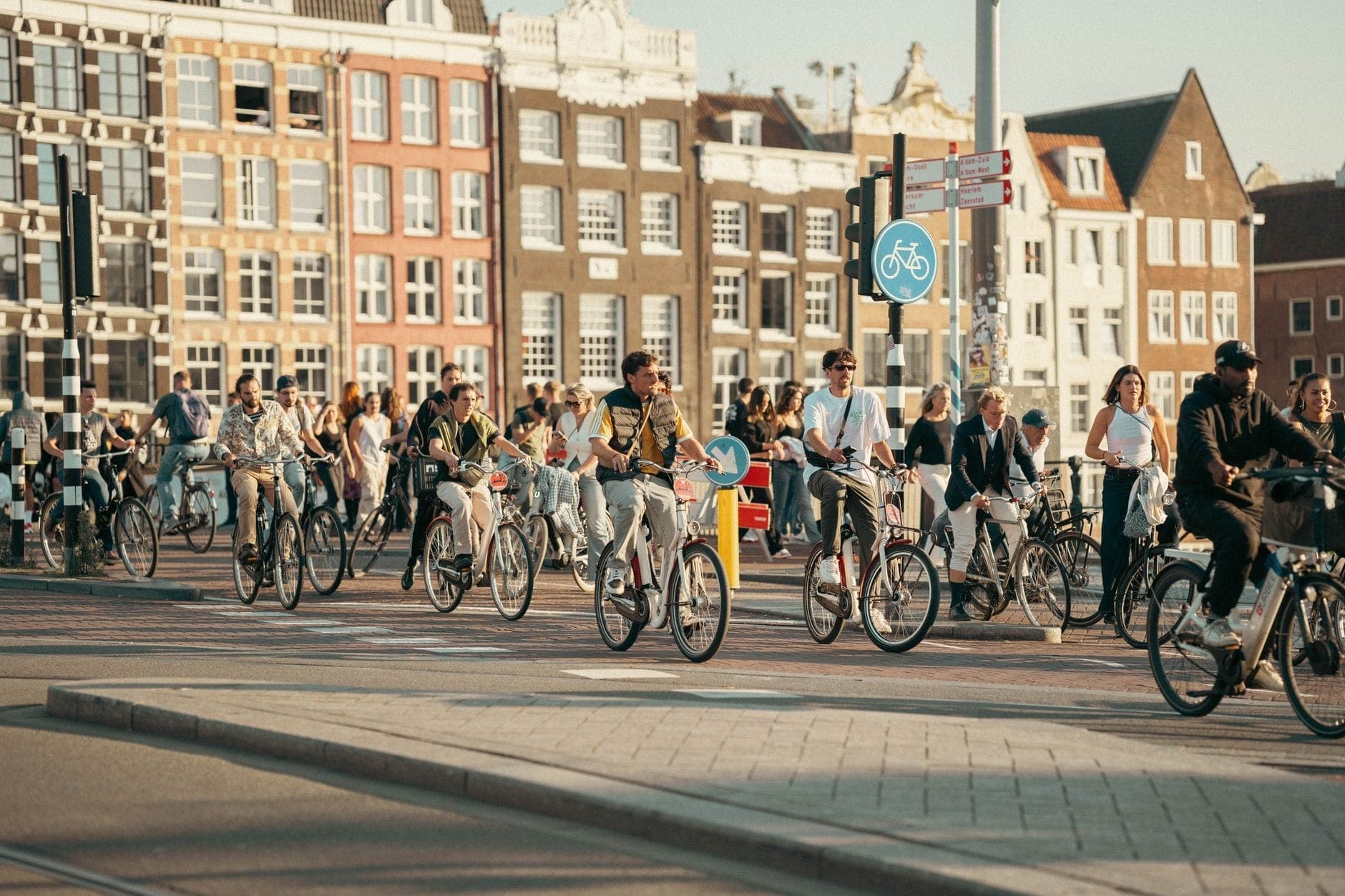 Eine große Gruppe von Menschen fährt an einem sonnigen Tag eine Amsterdamer Straße entlang, im Hintergrund typisch niederländische Grachtenhäuser.