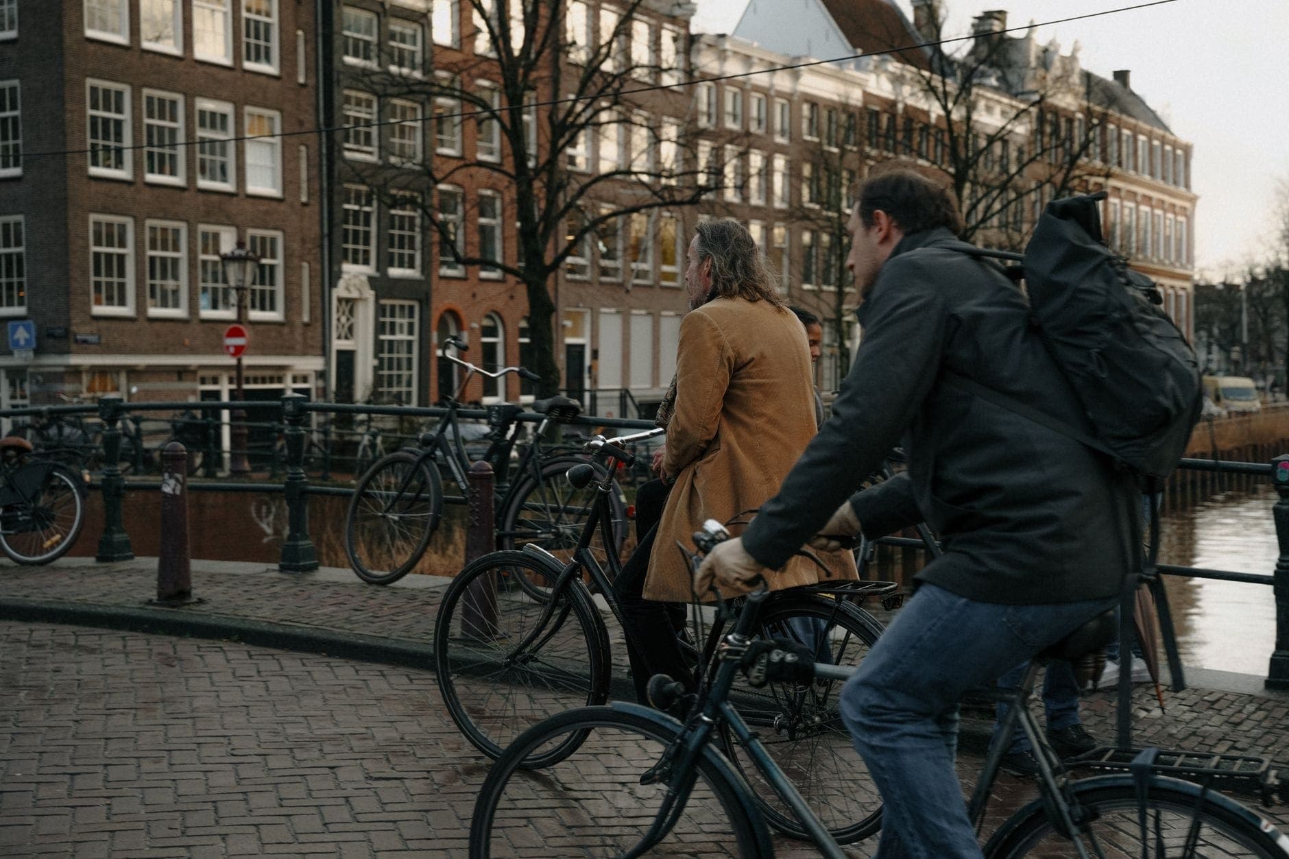 Zwei Radfahrer auf einer Brücke über einen Kanal in Amsterdam, umgeben von geparkten Fahrrädern und historischen Gebäuden am Kanalufer.