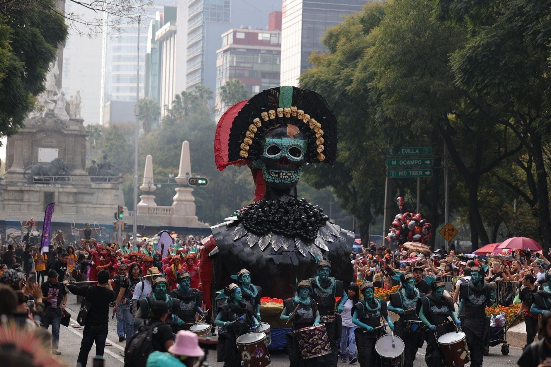 Défilé du Día de los Muertos à Mexico avec un char squelette élaboré et une grande foule sur une avenue, entourée de bâtiments et d'arbres.