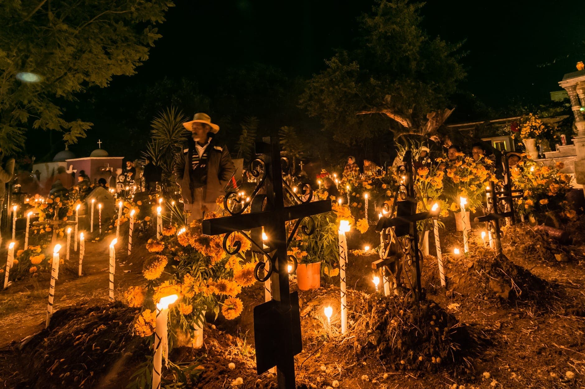 Scène nocturne dans un cimetière éclairé aux bougies avec des fleurs de souci, des croix et des personnes observant les traditions du Día de los Muertos.