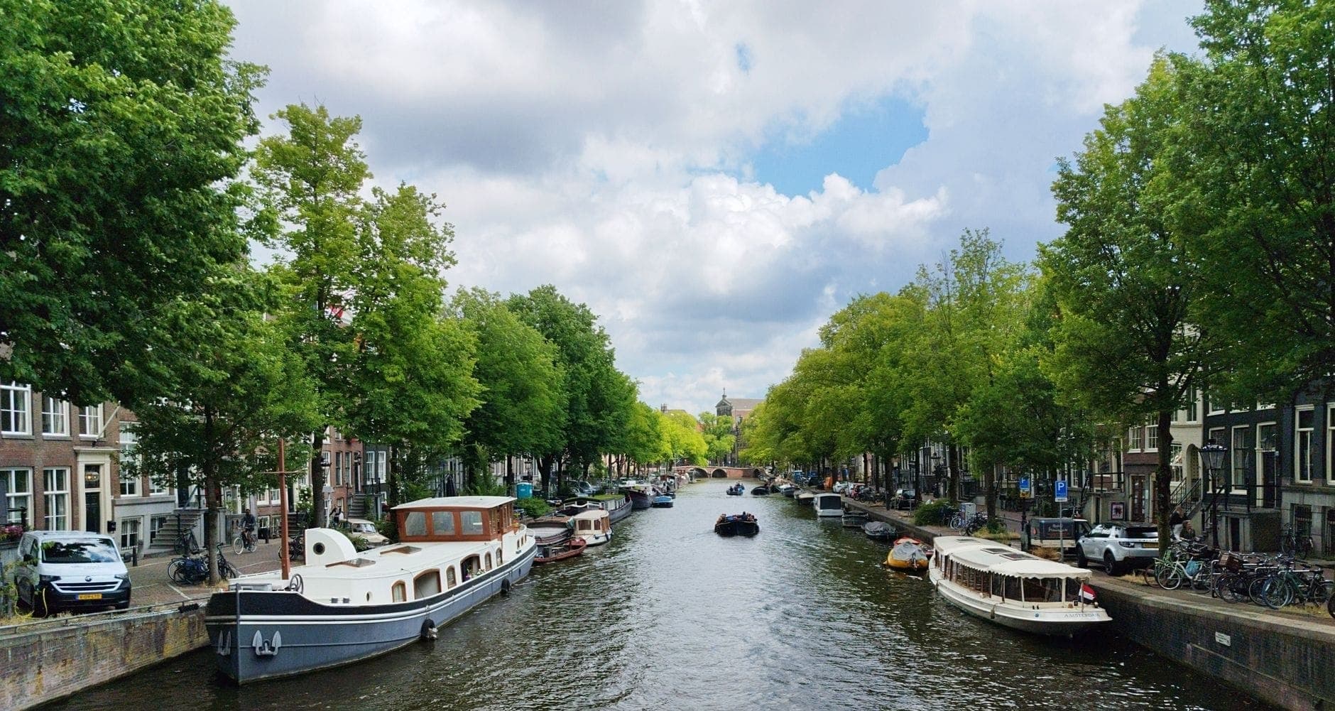 Weiter Blick auf einen malerischen Amsterdamer Kanal mit Bäumen und Hausbooten unter leicht bewölktem Himmel – einladend und bereit zum Entdecken.