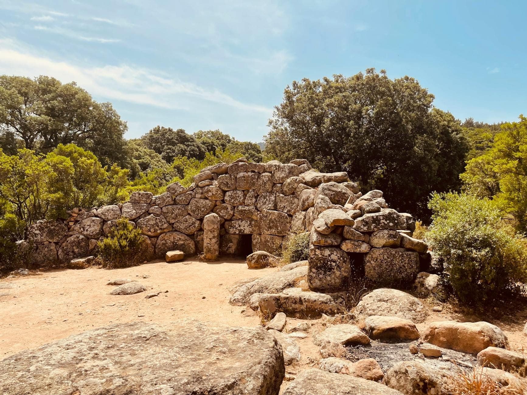 Antigua estructura nurágica de piedra entre árboles y rocas bajo un cielo azul en la Cerdeña rural.