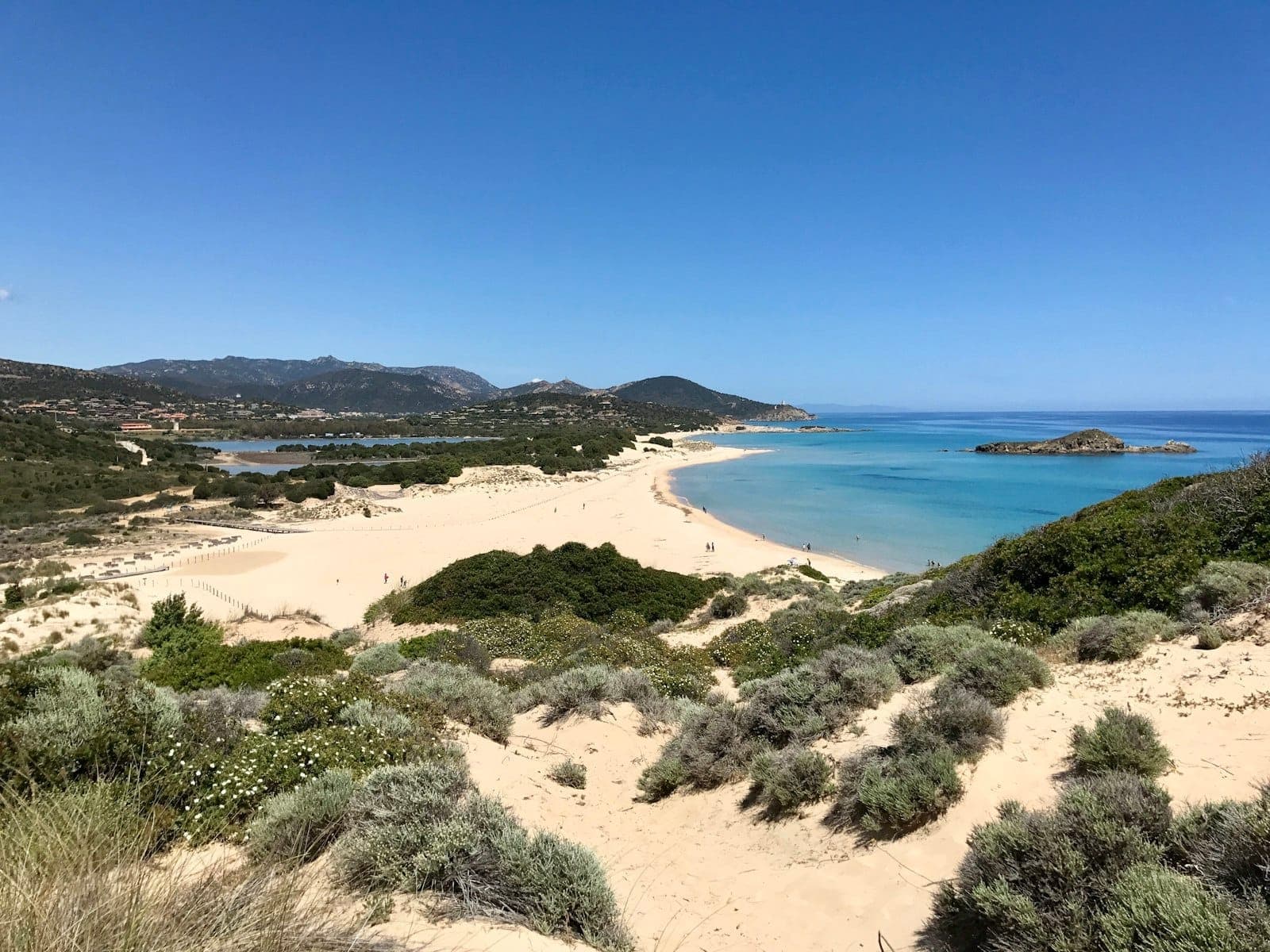 Vista amplia de una playa de arena prístina con agua turquesa, olas suaves y arbustos mediterráneos cerca de Cagliari, Cerdeña bajo un cielo azul despejado.