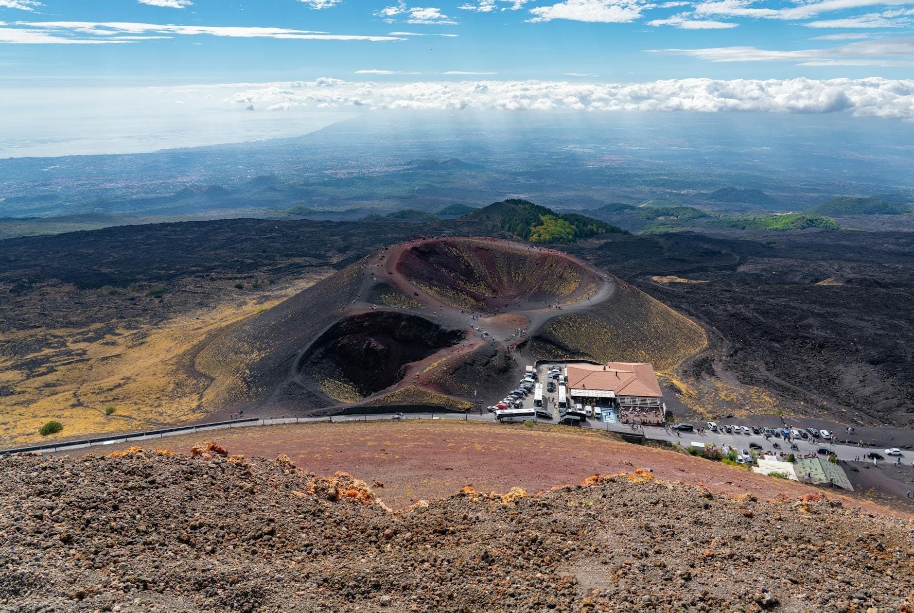 Pemandangan panoramik kawah vulkanik Gunung Etna dengan stasiun wisata dan mobil di dasarnya, berlatar garis pantai Sisilia yang jauh dan langit.