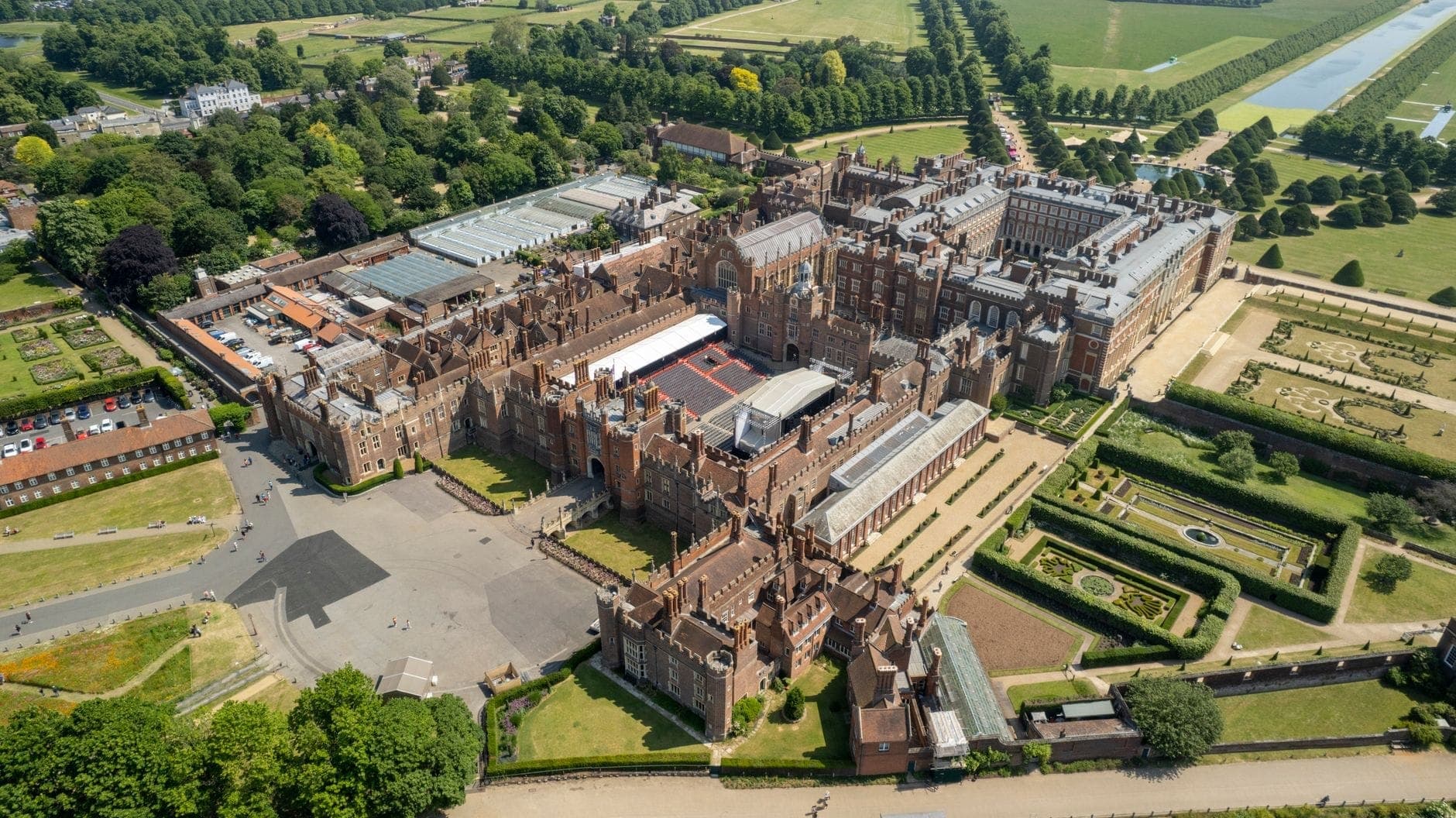 Luftaufnahme des Hampton Court Palace und seiner formalen Gärten, umgeben von Bäumen und Parklandschaft im Themse-Tal nahe London.