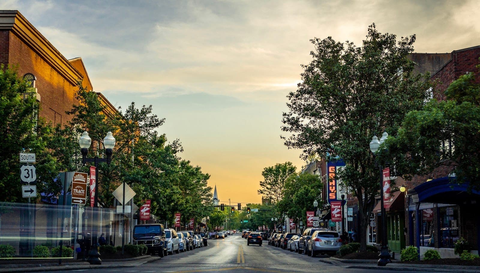 Incantevole strada principale di una piccola città fiancheggiata da alberi, edifici in mattoni, auto parcheggiate e striscioni al tramonto, che invita a un'esplorazione tranquilla.