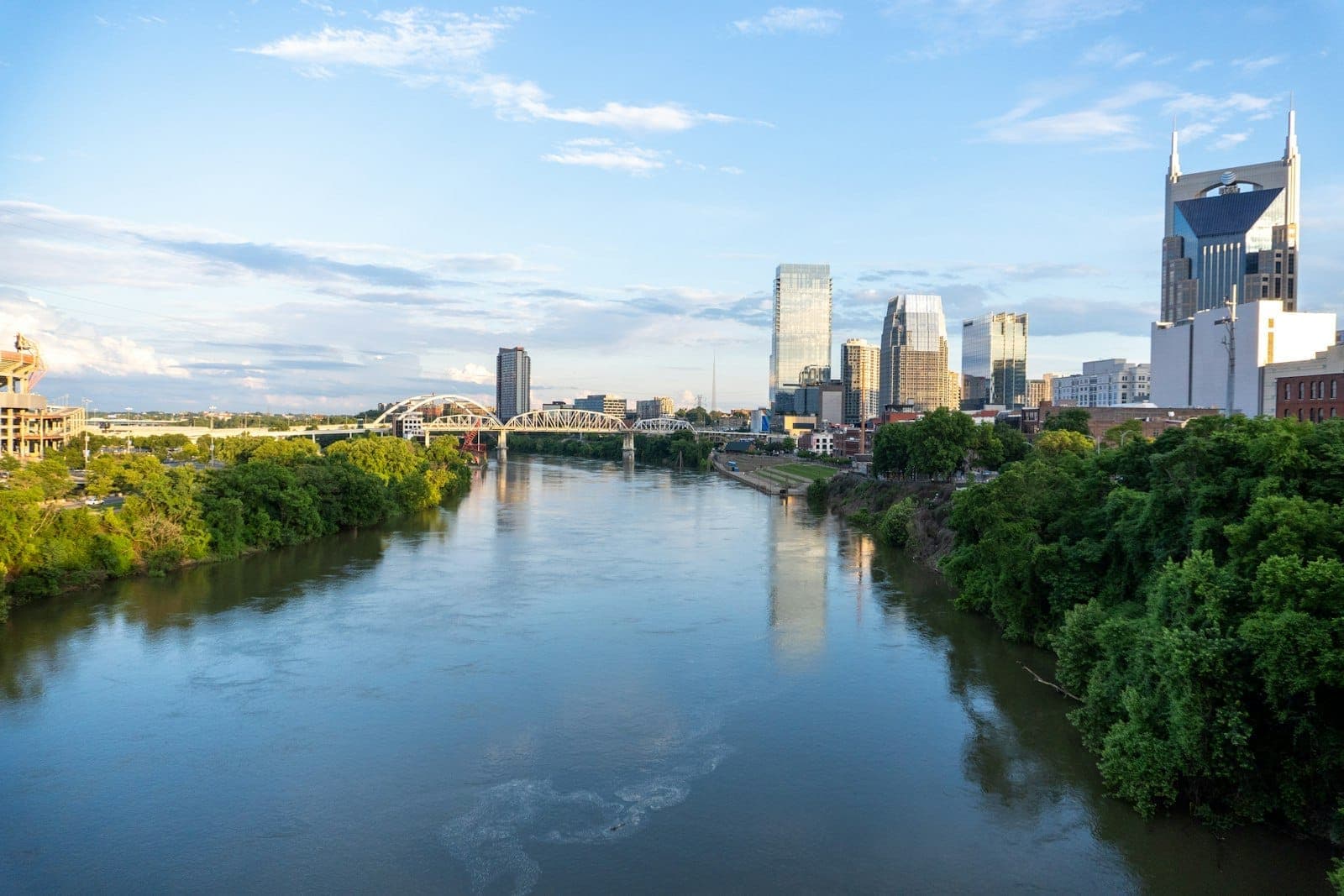Wide scenic view of the Nashville skyline behind a river, green trees along the water under a bright blue sky with soft clouds.