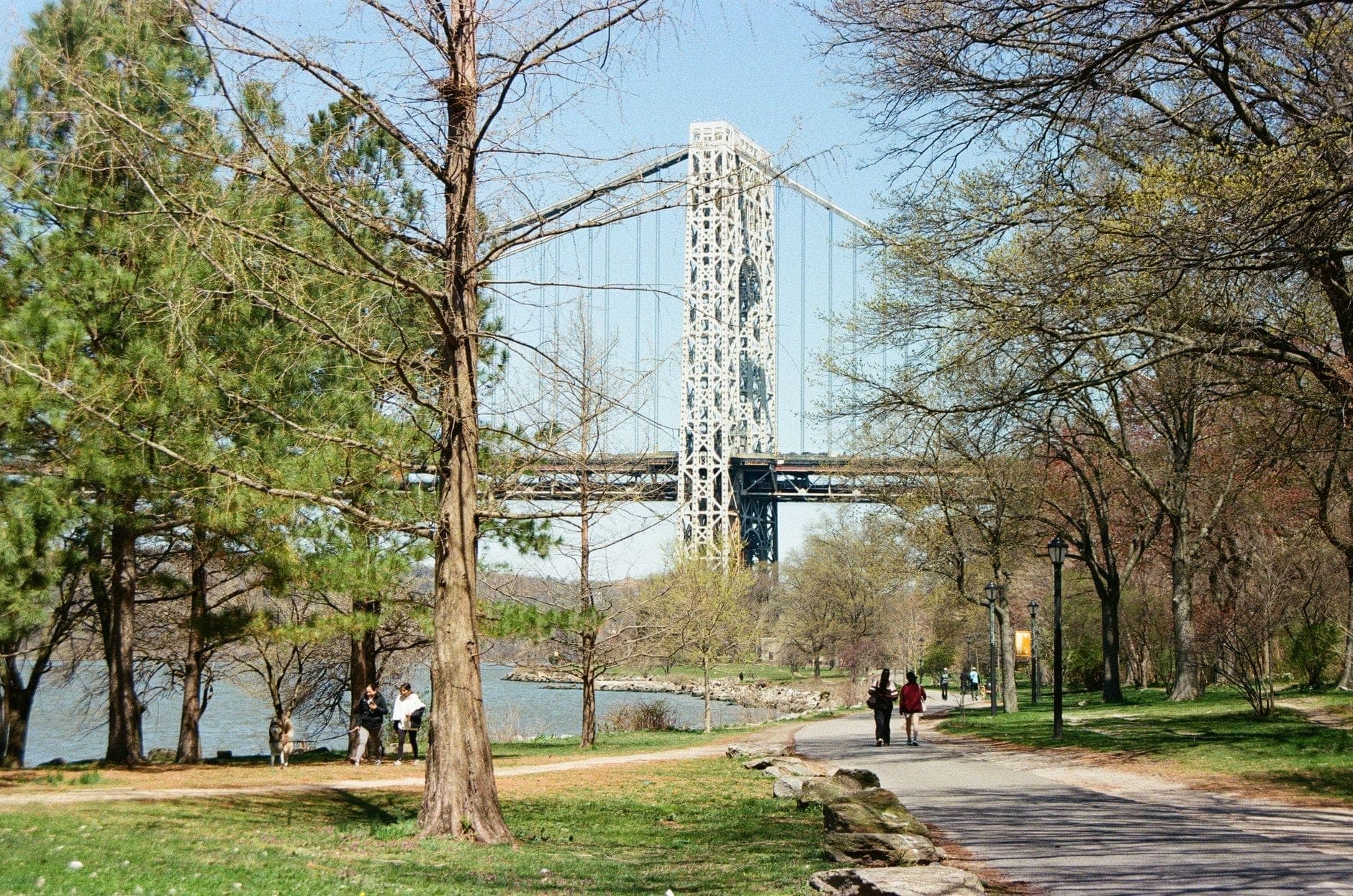 ทัศนียภาพสวนสาธารณะริมน้ำที่มีผู้คนเดินเล่น ต้นไม้ และ George Washington Bridge อยู่เบื้องหลังใน Upper Manhattan