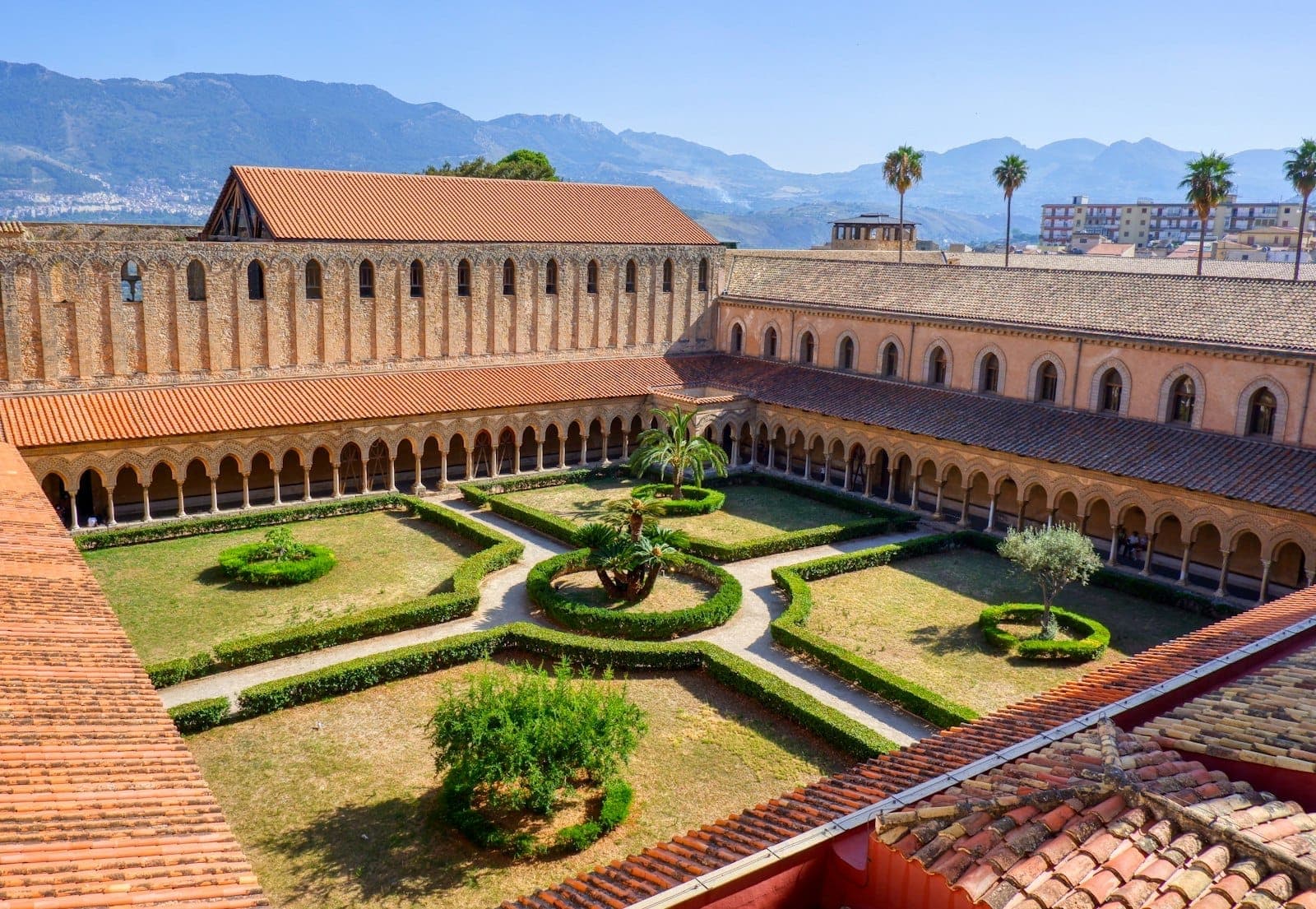 Pemandangan udara serambi Katedral Monreale dengan kolom-kolom yang rumit, jalur taman, dan lanskap pegunungan yang jauh di bawah langit biru cerah.