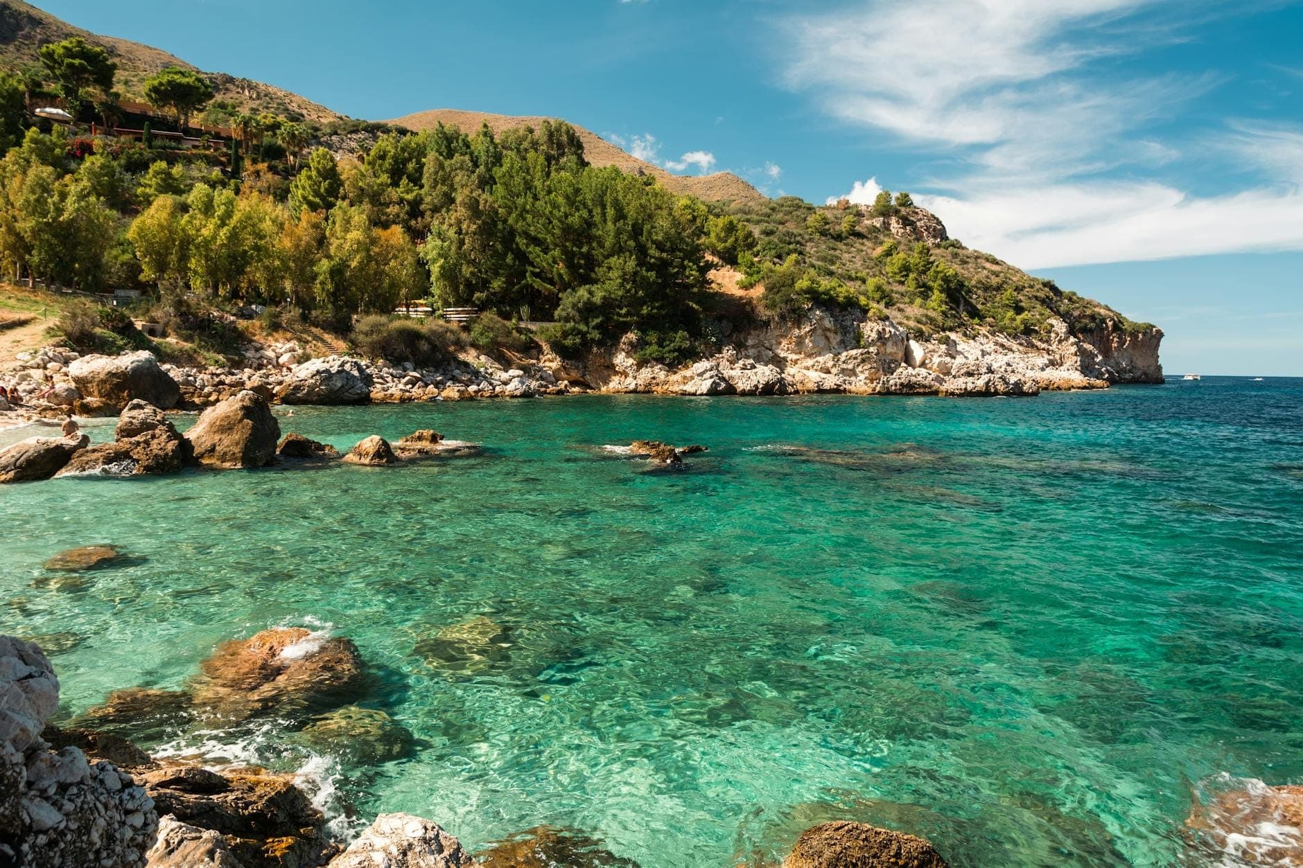 Pantai berbatu dengan air pirus jernih dan pohon-pohon hijau lebat di bawah langit biru, khas dari cagar alam Sisilia.