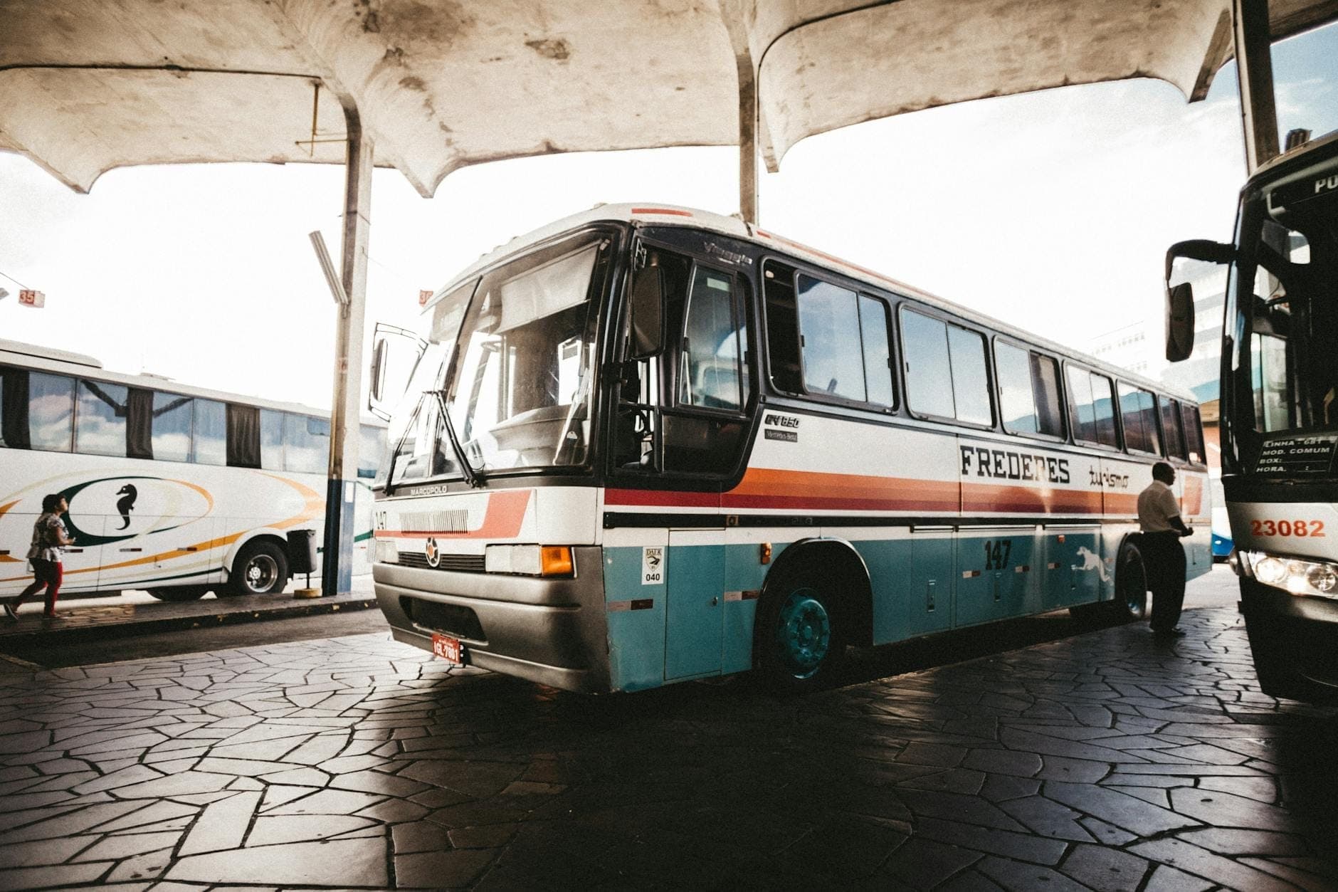Un autobús clásico azul y blanco estacionado en una terminal cubierta, con pasajeros cercanos, que sugiere el transporte público típico de las islas griegas.