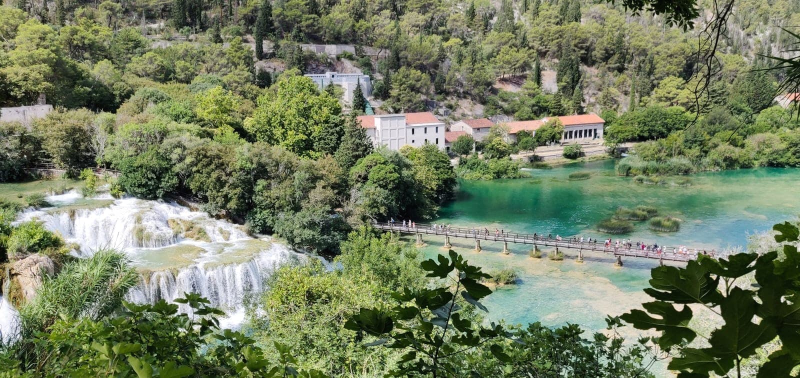 Vista panoramica del Parco Nazionale di Krka con le famose cascate di Skradinski Buk, il fiume turchese, antichi edifici in pietra e un ponte in legno circondato da una lussureggiante vegetazione.