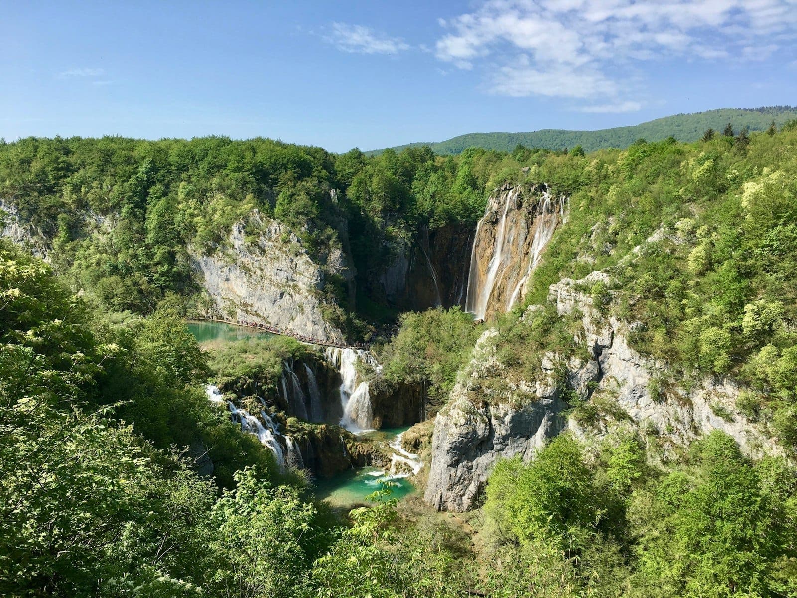 Vue des cascades spectaculaires et des bassins turquoise des Lacs de Plitvice entourés d'une forêt verdoyante luxuriante et de falaises rocheuses sous un ciel bleu.
