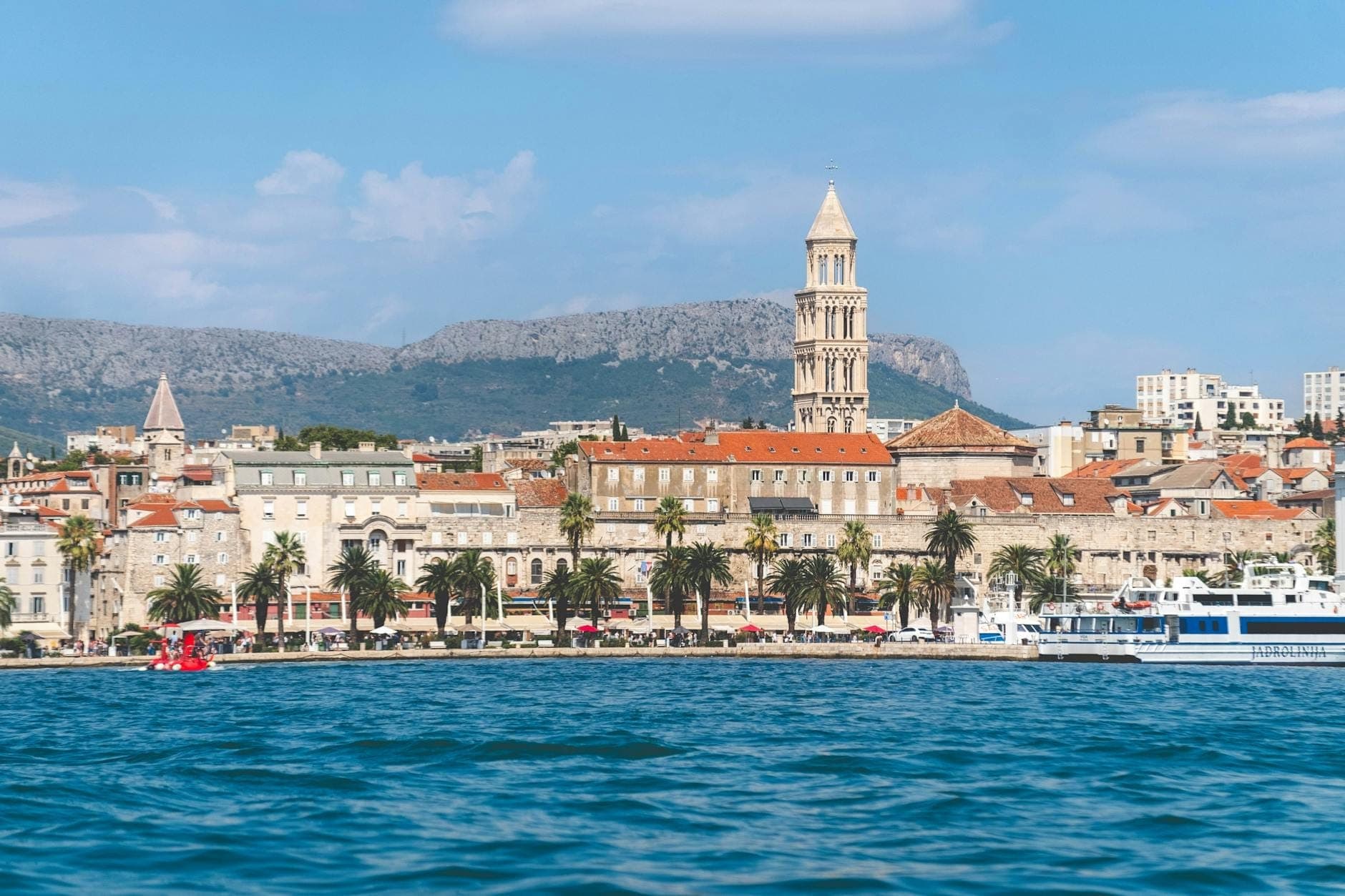 Vista panoramica del lungomare di Spalato con palme, barche, il mare Adriatico e l'antico campanile sullo sfondo delle montagne, sotto un cielo azzurro e luminoso.