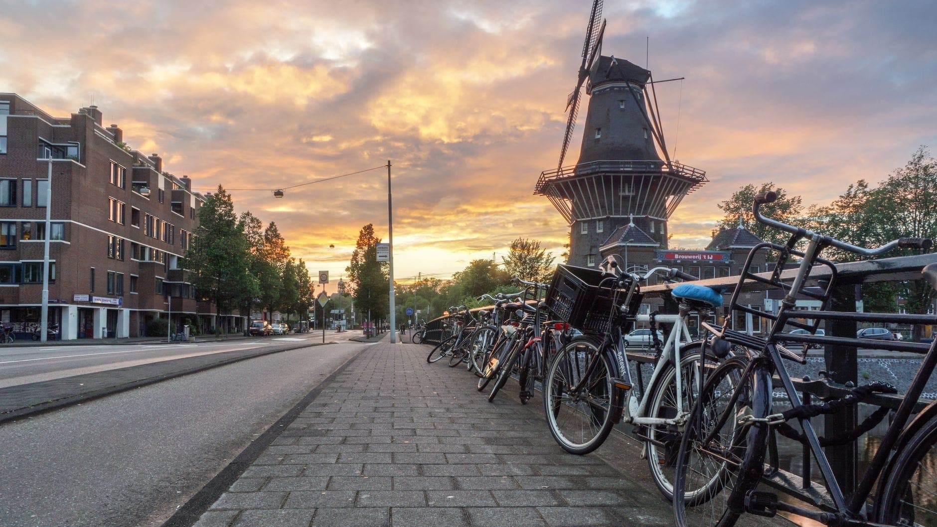 Amsterdam street scene at sunset with De Gooyer windmill in the background, lined with bicycles and historic buildings, evoking urban Dutch charm.