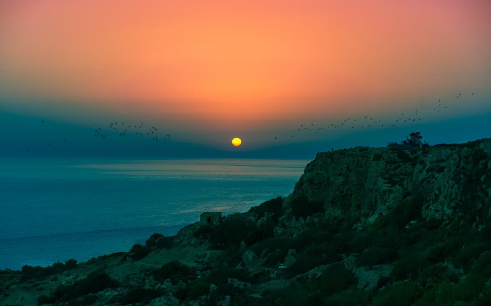 Dramatic sunset over Dingli Cliffs with the sun setting above the sea, silhouetted cliff edges, and a glowing colorful sky.