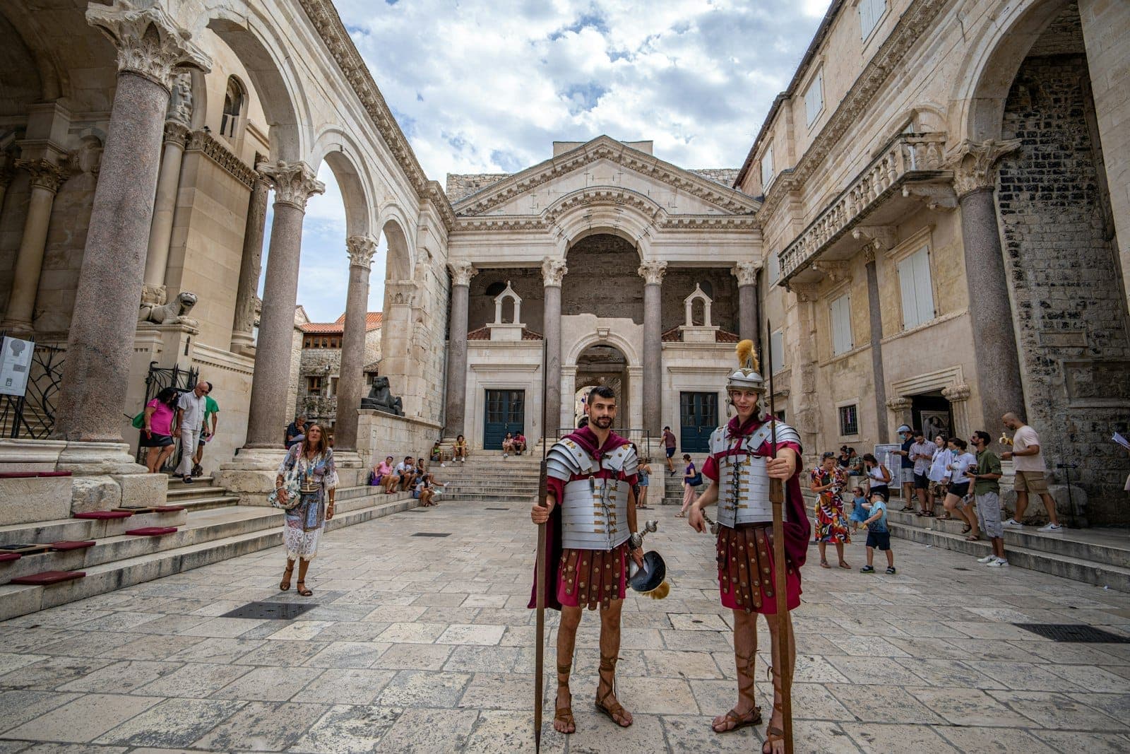 Visitatori e attori in costumi romani si riuniscono nel cortile del Peristilio del Palazzo di Diocleziano, circondati da grandi colonne e archi in pietra calcarea antica.