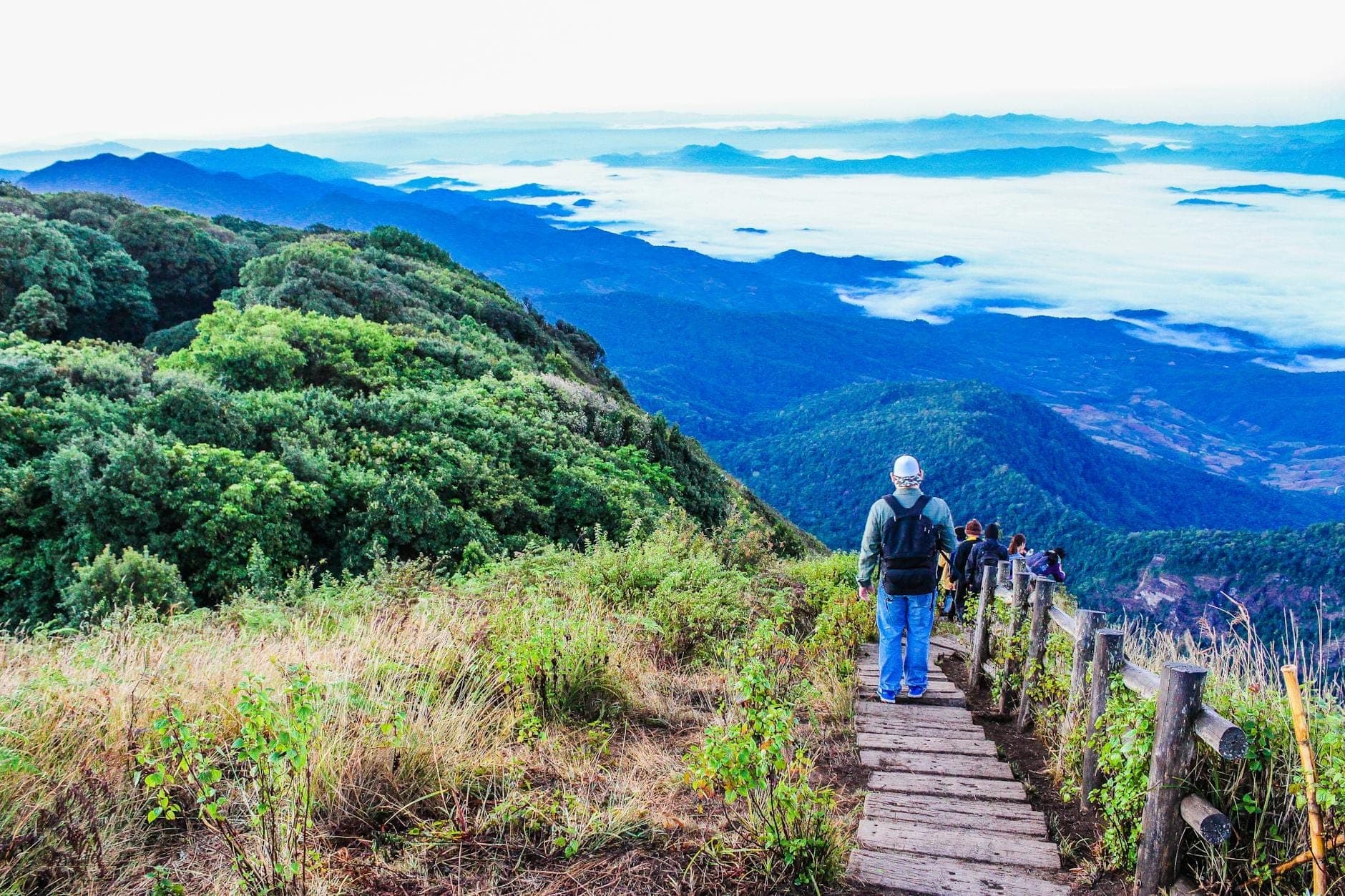 Travelers walking along a wooden path on a scenic mountain ridge with lush forest and a cloud-filled valley below.