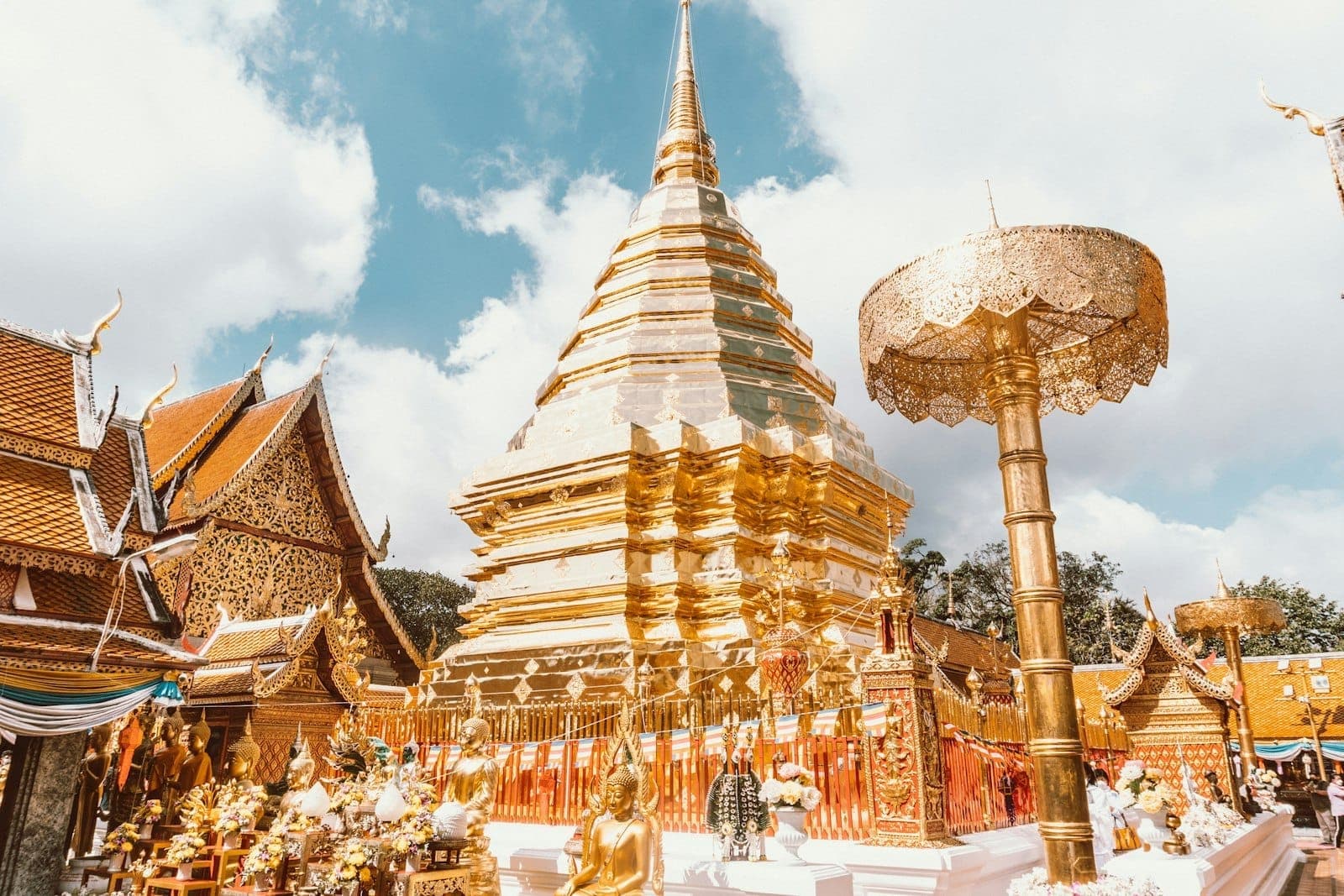 Der leuchtend goldene Stupa des Wat Phra That Doi Suthep vor blauem Himmel mit Wolken, umgeben von kunstvollen Bauwerken und Besuchern im Berggebiet von Chiang Mai.