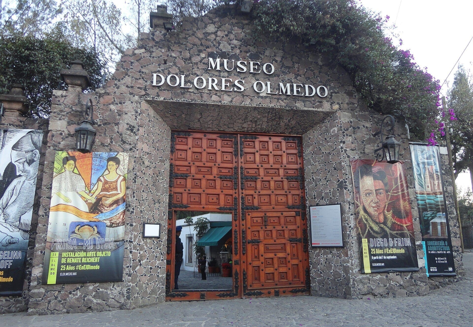 ทางเข้าหินของ Museo Dolores Olmedo ในเม็กซิโกซิตี้ มีประตูไม้ขนาดใหญ่ ป้ายแบนเนอร์หลากสี และพืชพรรณเขียวชอุ่มเหนือประตูทางเข้า