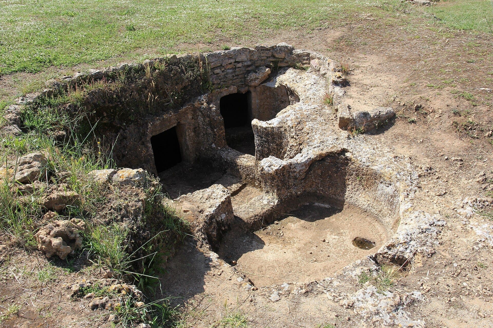 Vista de las antiguas tumbas excavadas en la roca en Domus de Janas di Anghelu Ruju, con cámaras de arenisca expuestas en una zona abierta con hierba.