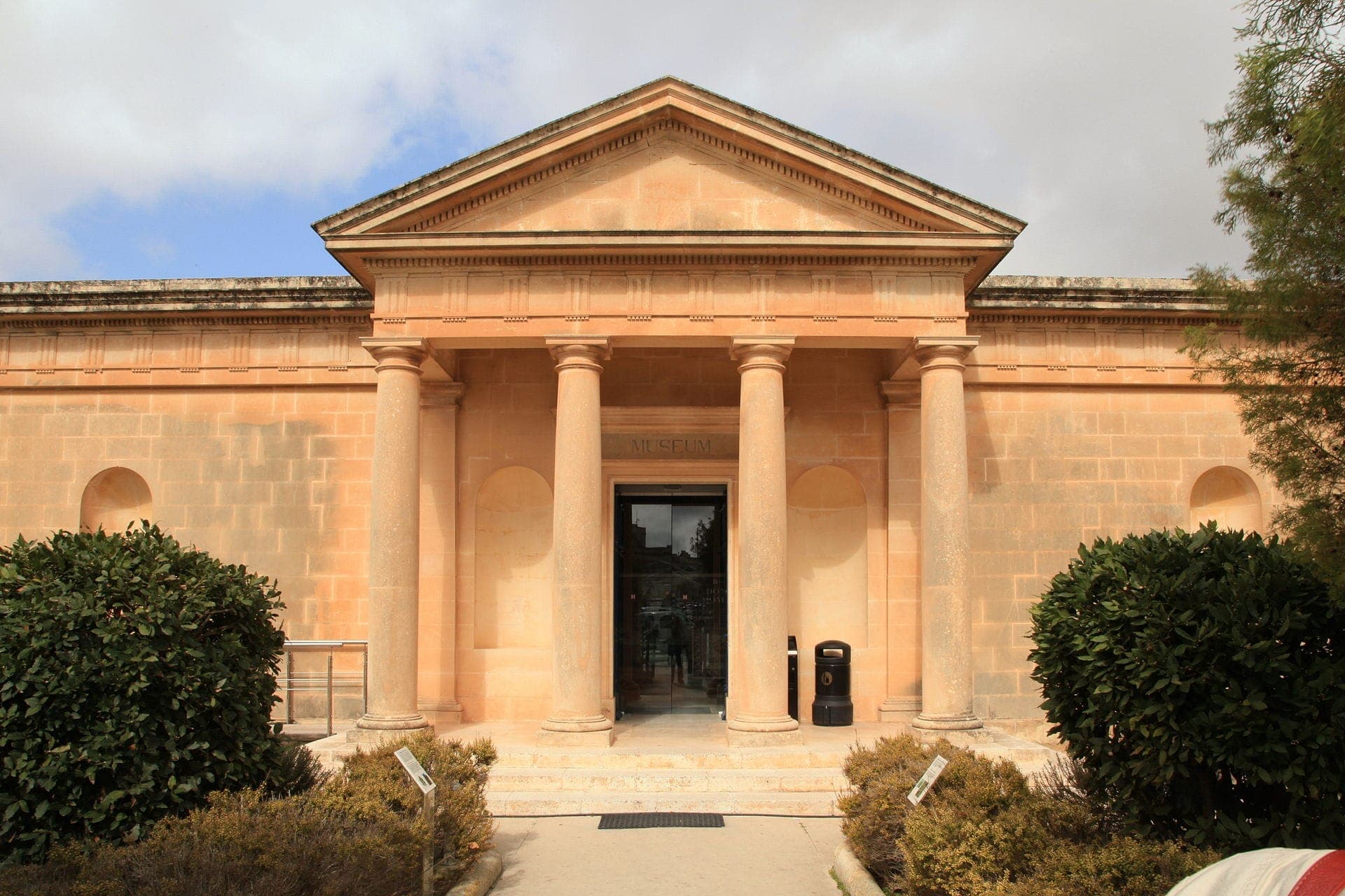 Frontansicht des Museums Domus Romana in Malta mit neoklassischen Säulen, sandfarbenem Stein und gepflegten Sträuchern unter einem teilweise bewölkten Himmel.