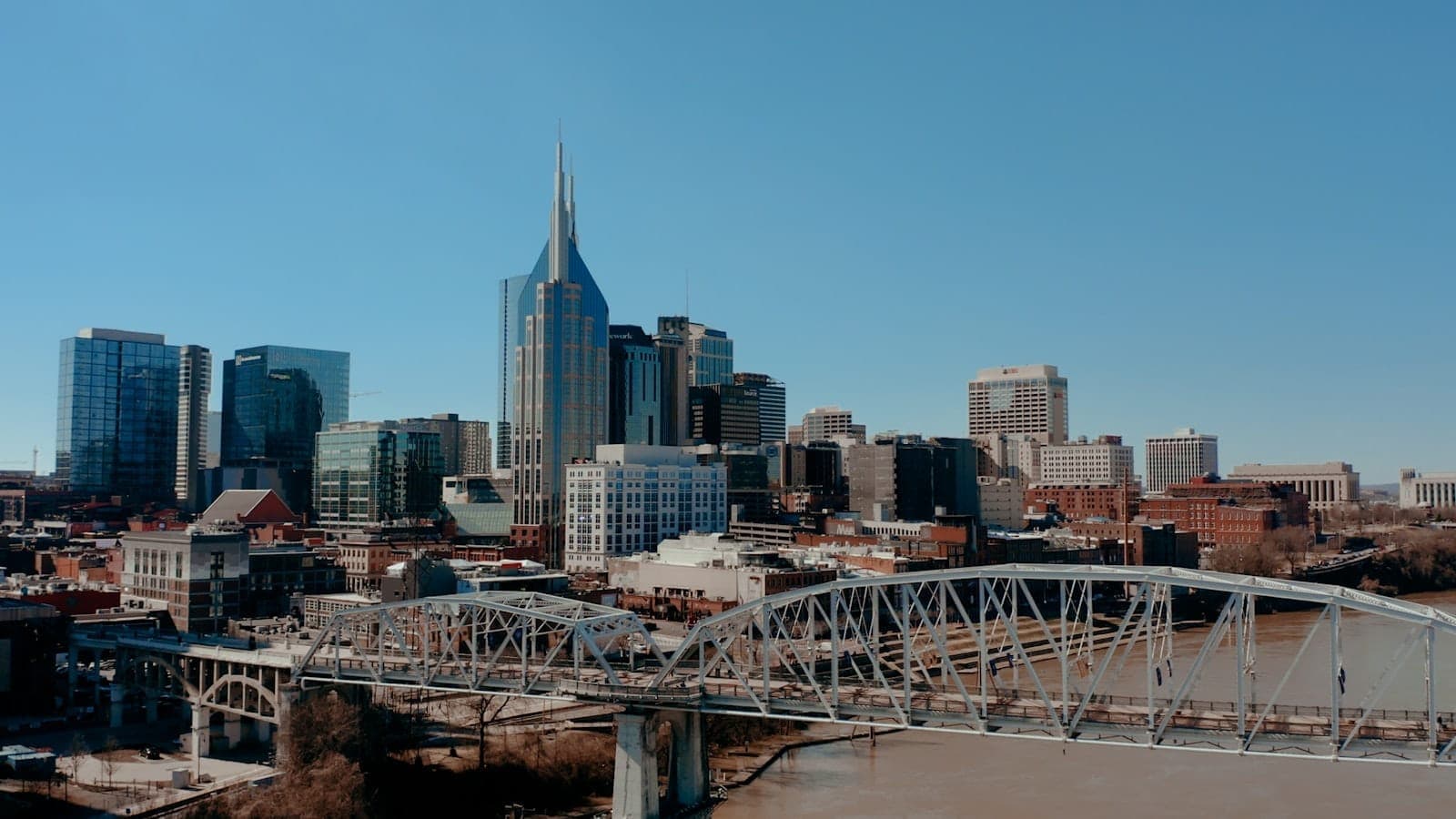 Wide view of downtown Nashville skyline with modern skyscrapers, historic buildings, and a bridge crossing the Cumberland River on a clear sunny day.