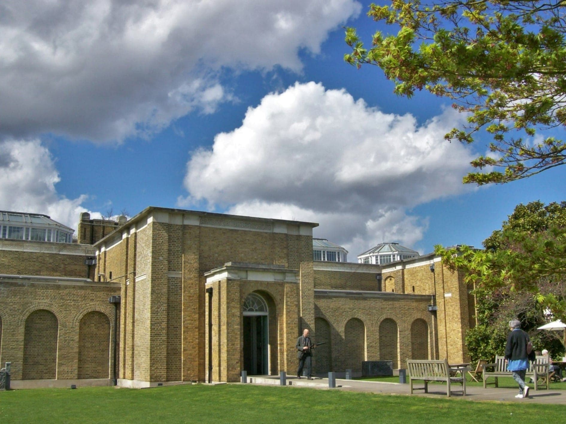Die stattliche gelbe Backsteinfront der Dulwich Picture Gallery unter einem dramatisch bewölkten Himmel, mit Bänken und Besuchern im Vordergrund.