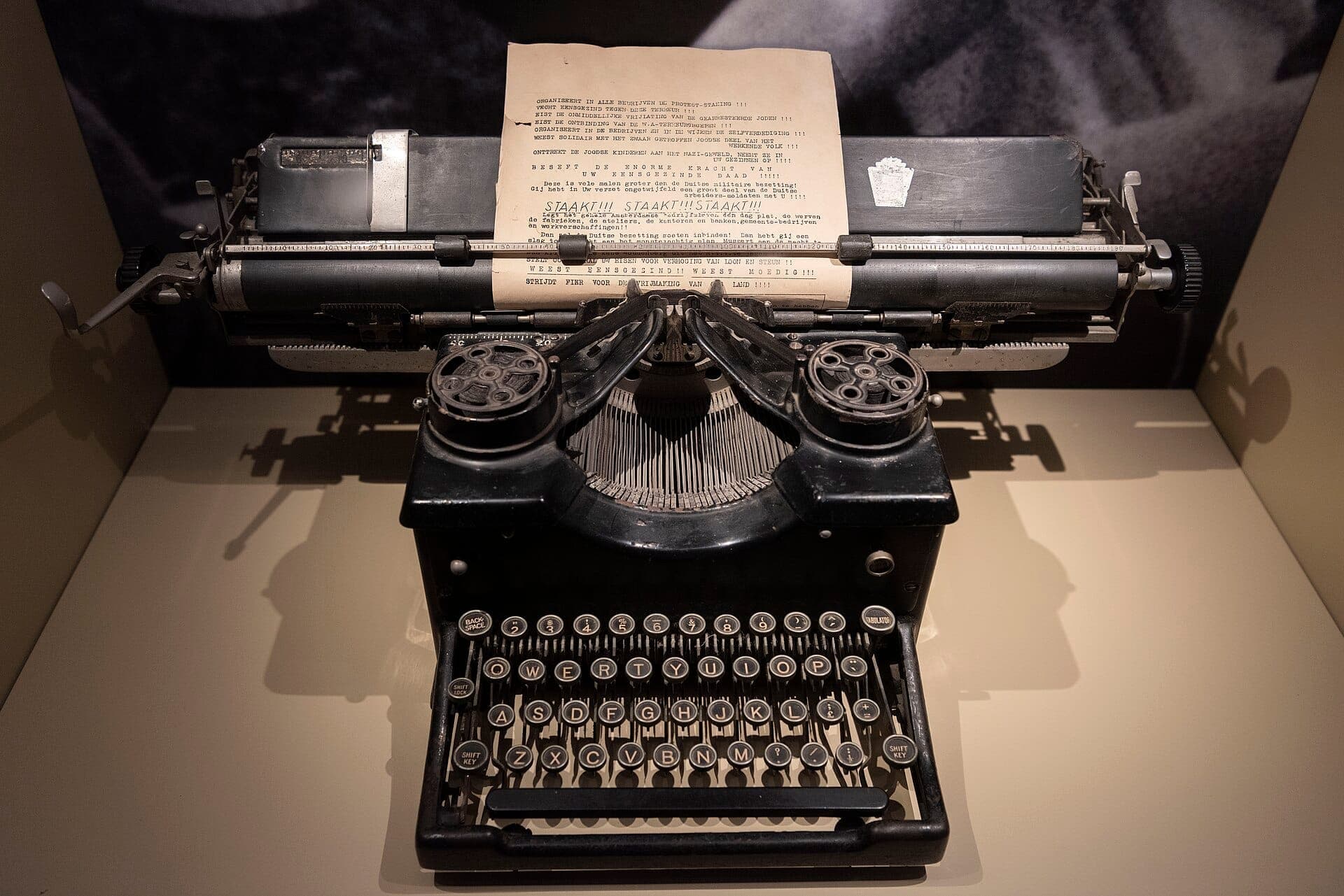 Vintage schwarze Schreibmaschine mit einem getippten Widerstandsdokument, ausgestellt im Dutch Resistance Museum in Amsterdam, gut beleuchtet vor einem neutralen Hintergrund.