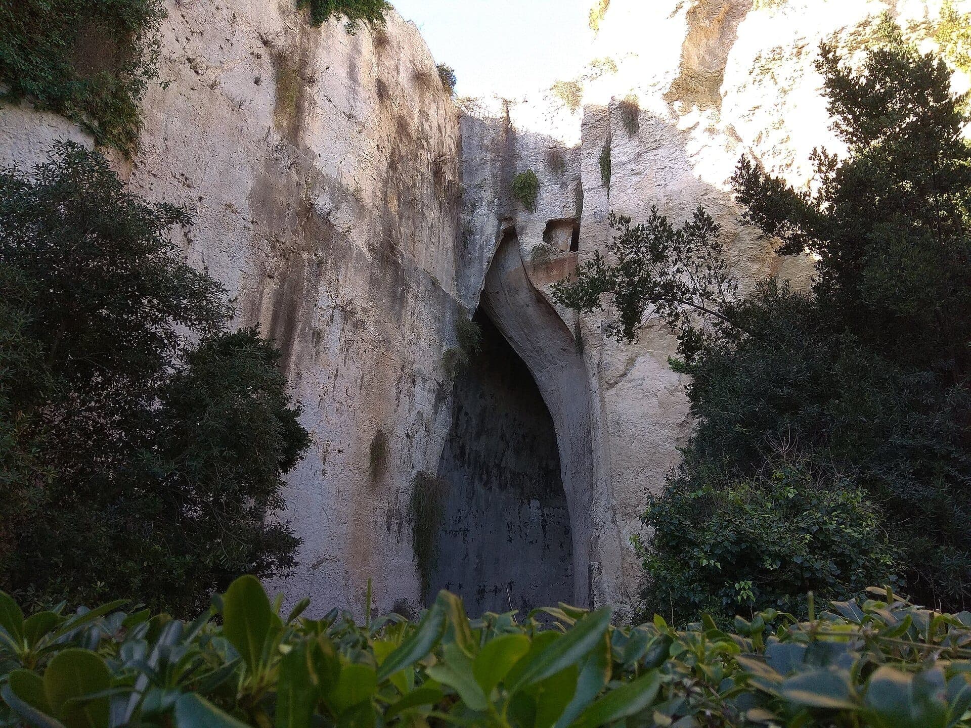 Pintu masuk Telinga Dionysius, sebuah gua batu kapur yang tinggi dikelilingi pepohonan hijau subur dan dinding batu yang lapuk di Syracuse, Sisilia.