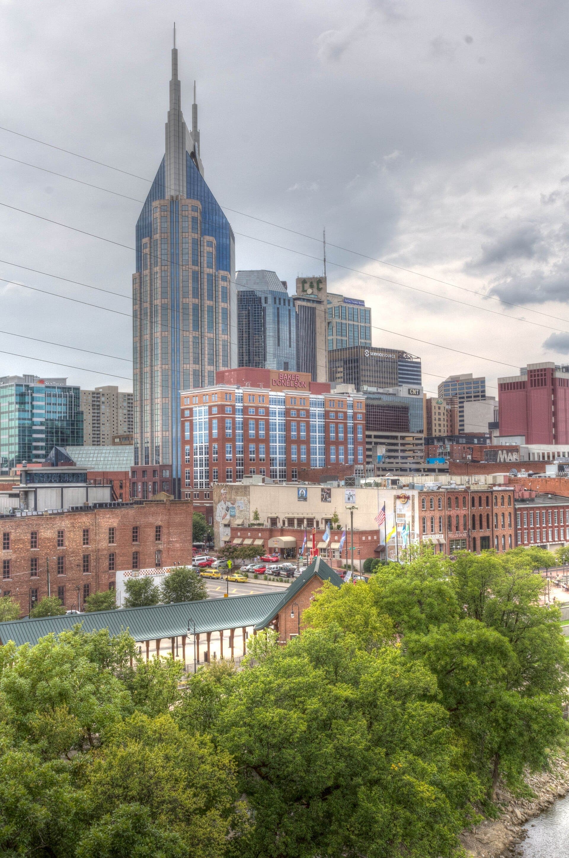 Downtown Nashville skyline seen from East Nashville, featuring the iconic AT&T Building, historic brick buildings, and lush green trees in the foreground.