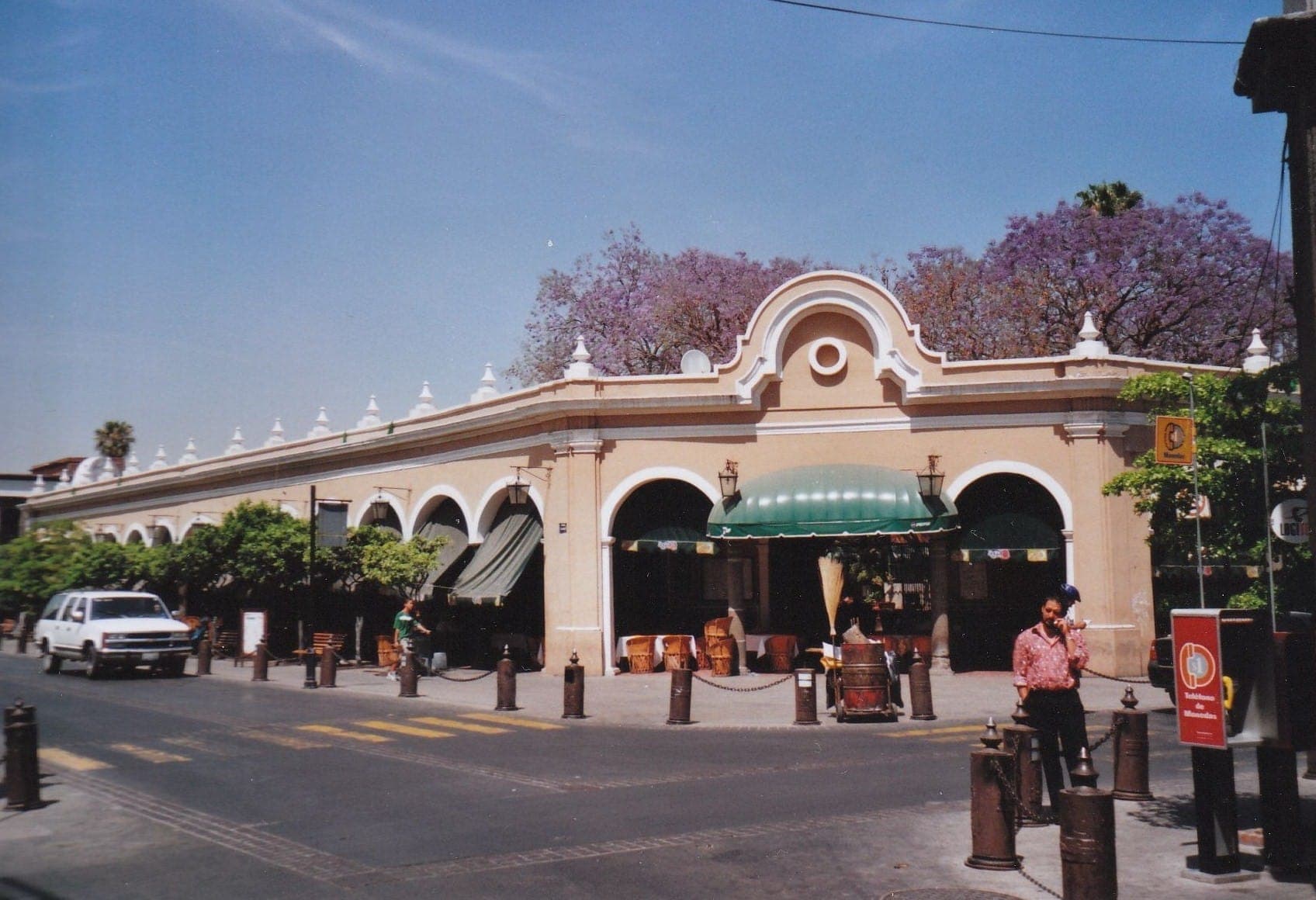 El Parián de Tlaquepaque อันเก่าแก่ที่มีด้านหน้าอาคารรูปโค้งและหลังคากันแดดสีเขียว ล้อมรอบด้วยต้นจาคารันดาที่กำลังออกดอกใต้ท้องฟ้าสีฟ้าใส