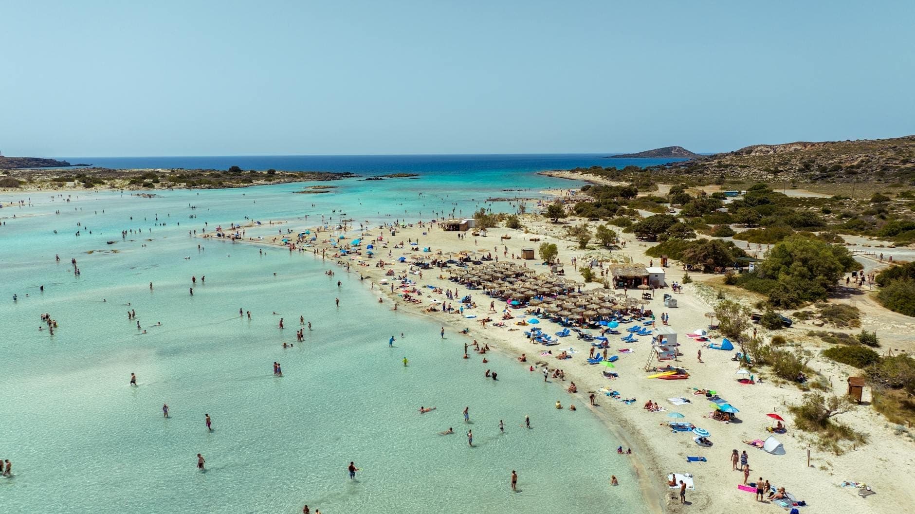 Luftaufnahme des Elafonissi Beach mit türkisfarbener Lagune, Menschenmassen auf rosafarbenem Sand, Sonnenschirmen und der kleinen Insel in der Ferne unter strahlend blauem Himmel.