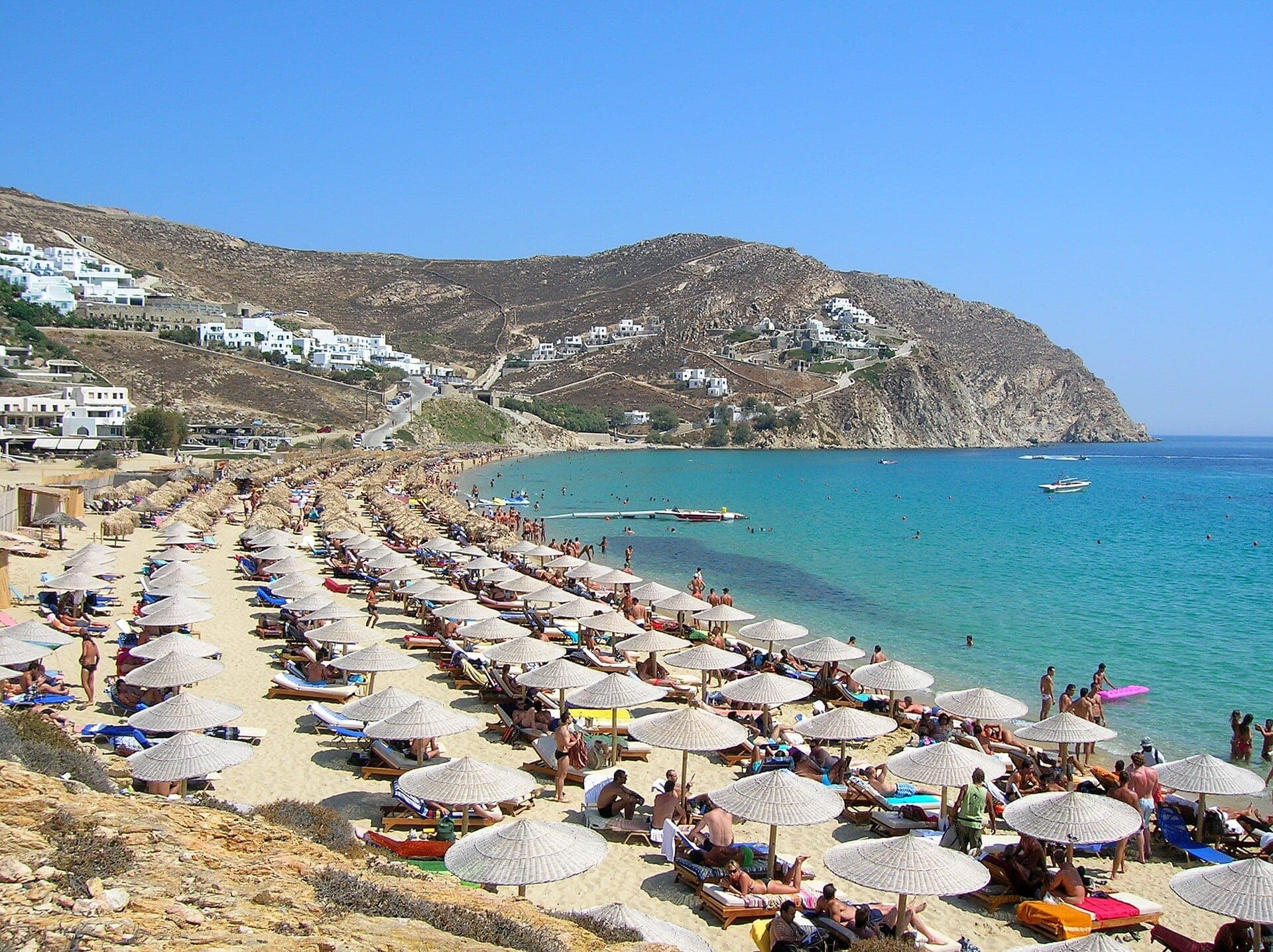 Reihen von Liegestühlen und Sonnenschirmen säumen den Elia Beach auf Mykonos, mit türkisfarbenem Wasser und weißen Gebäuden am Hang unter einem klaren blauen Himmel.
