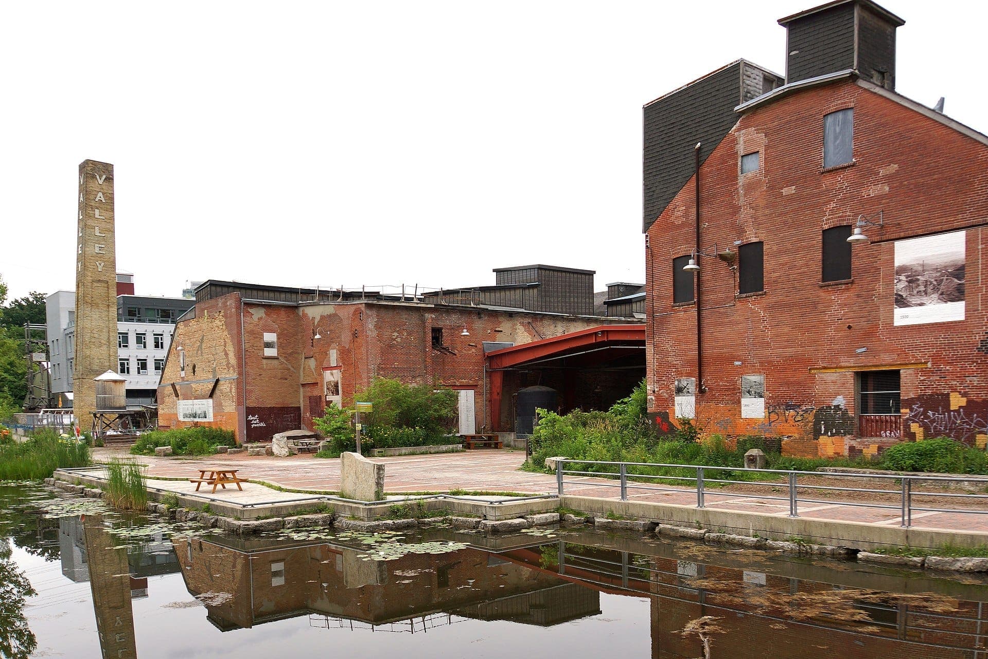 Edifici industriali in mattoni rossi e un alto camino si riflettono in uno stagno tranquillo all'Evergreen Brick Works in una giornata nuvolosa.