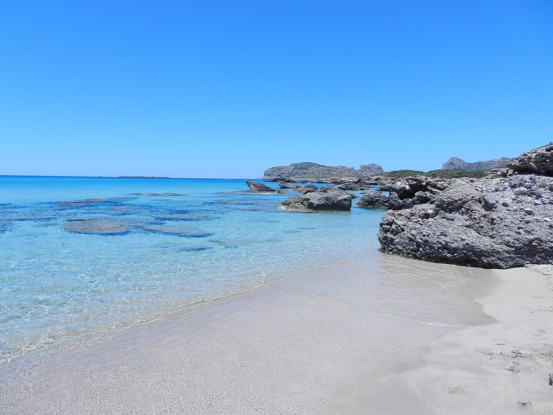 Klares türkisfarbenes Wasser und heller Sand am Falassarna Beach, Kreta, mit vereinzelten Felsvorsprüngen und fernen Hügeln unter einem strahlend blauen Himmel.