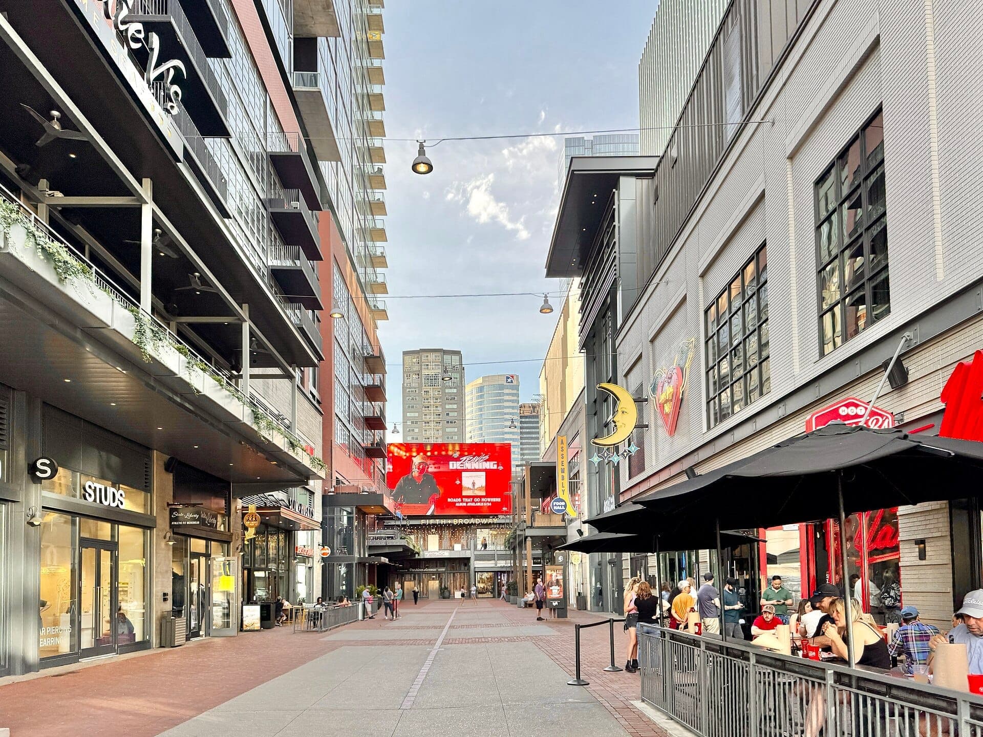 Street-level view of Fifth + Broadway in Nashville with modern buildings, glass storefronts, outdoor dining, and a digital billboard under a blue sky.