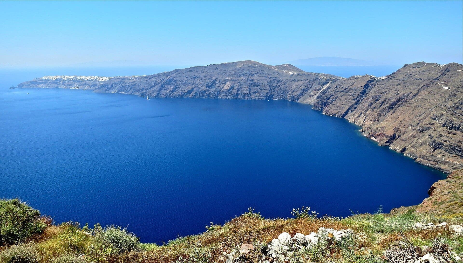 Blick auf die dramatischen Kaldera-Klippen von Santorini und das tiefblaue Ägäische Meer vom Wanderweg von Fira nach Oia, mit weißen Hangdörfern in der Ferne.