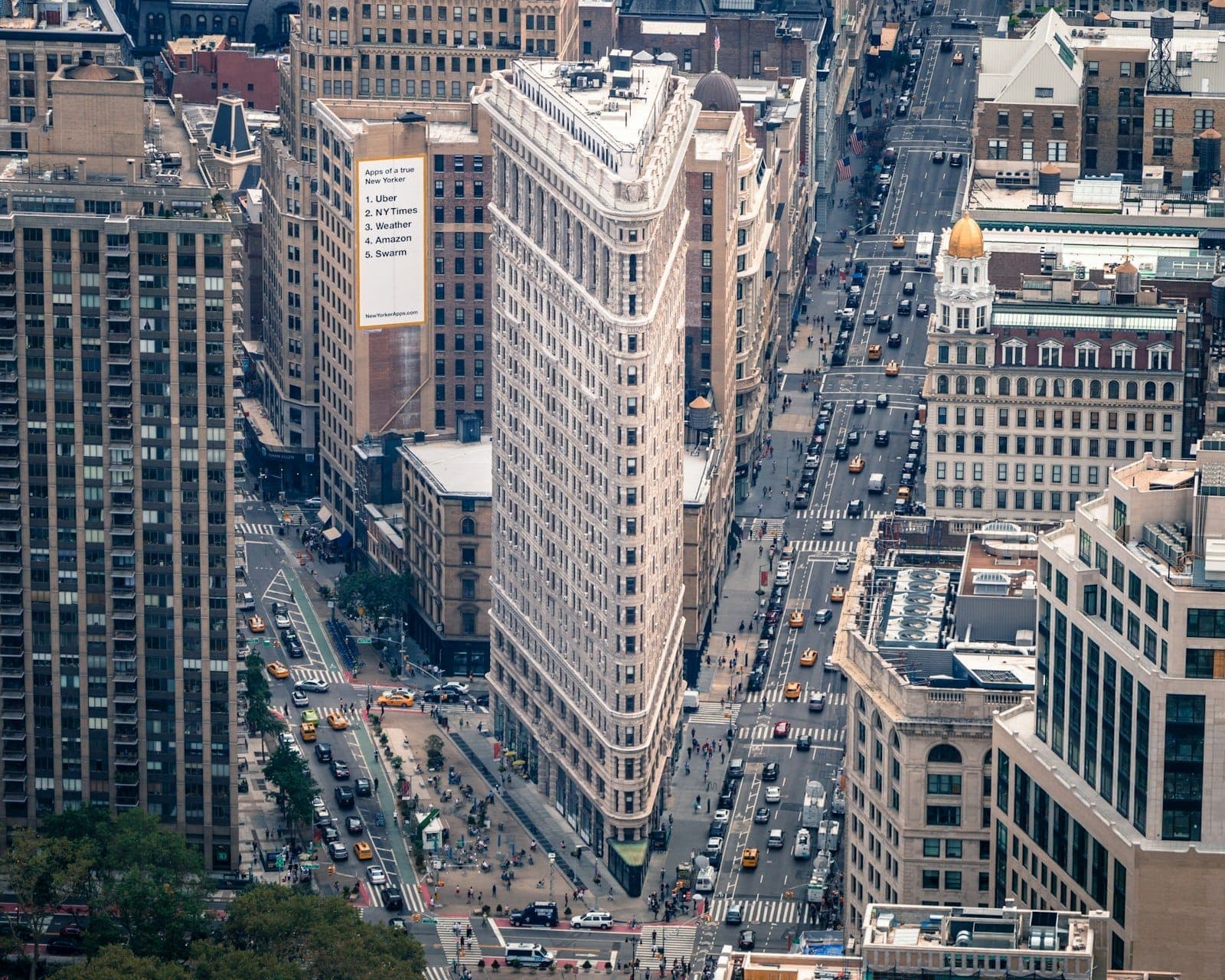 ภาพมุมสูงของอาคาร Flatiron เผยโครงสร้างสามเหลี่ยมแปลกตา รายล้อมด้วยถนนแมนฮัตตันอันวุ่นวาย รถแท็กซี่สีเหลือง และตึกเก่าใกล้เคียง
