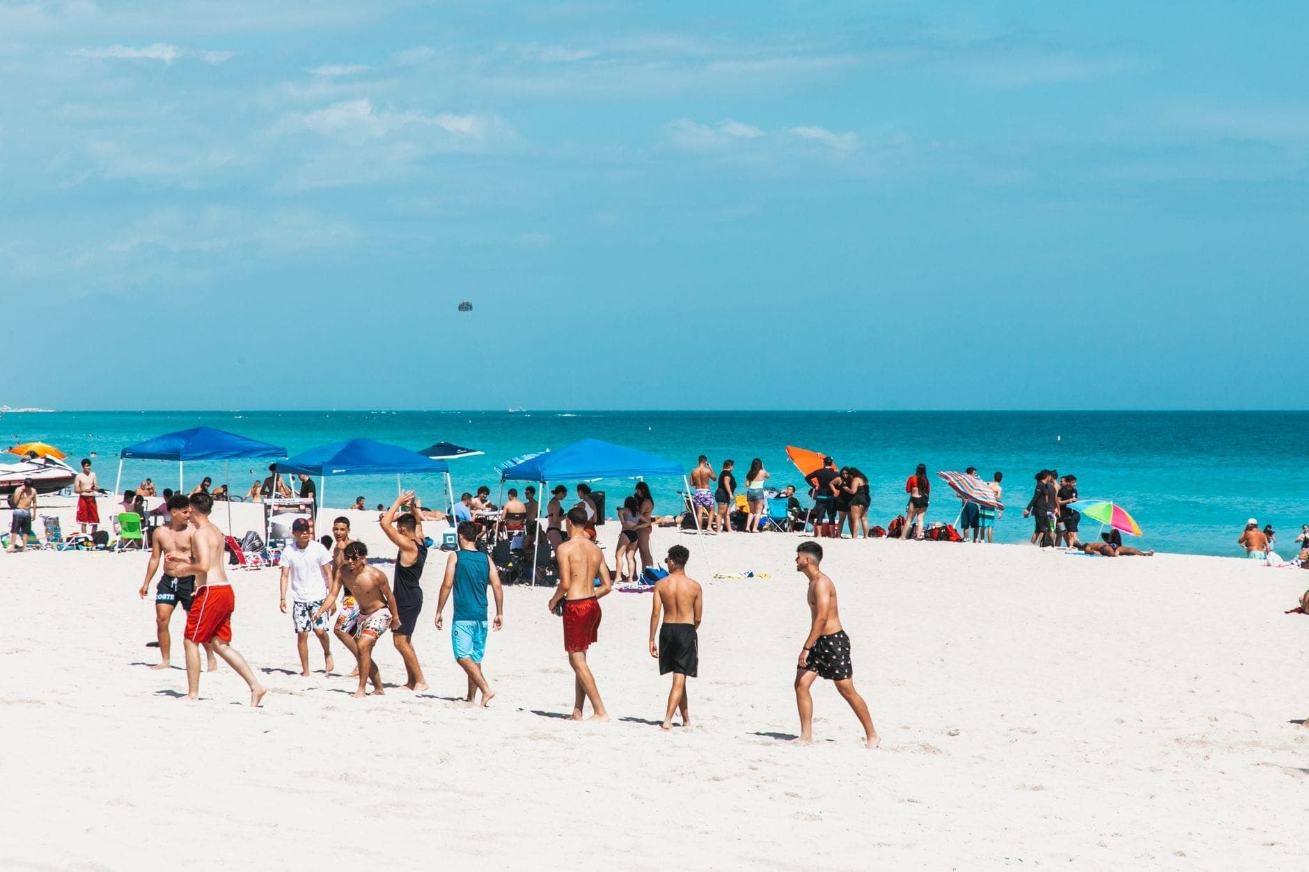 Crowded Miami beach with groups of people under colorful umbrellas, blue sky, and turquoise water on a sunny day.