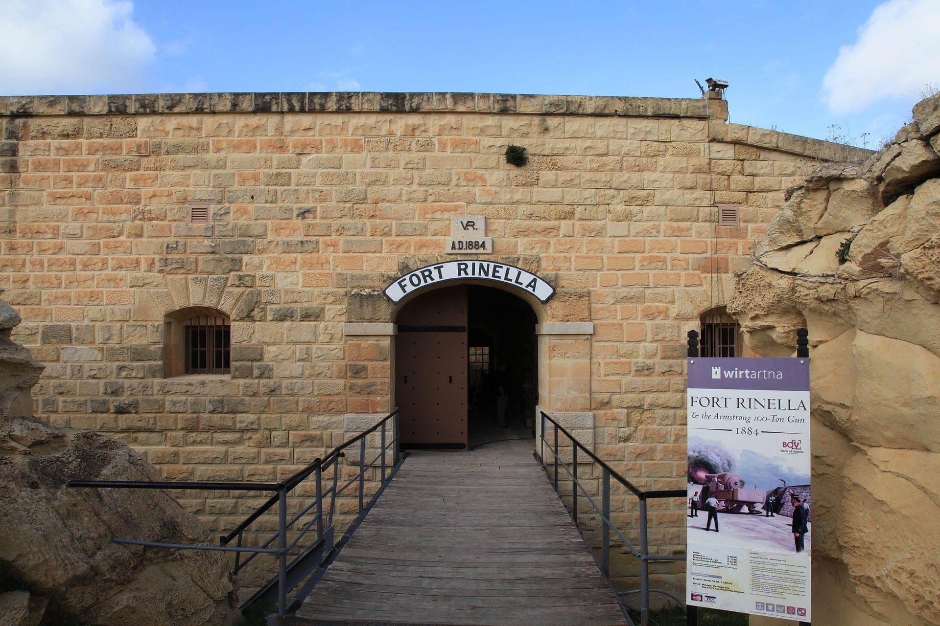 Eingang von Fort Rinella mit Sandsteinmauern, gewölbtem Eingangstor und einem Informationsschild unter einem teilweise bewölkten blauen Himmel in Kalkara, Malta.