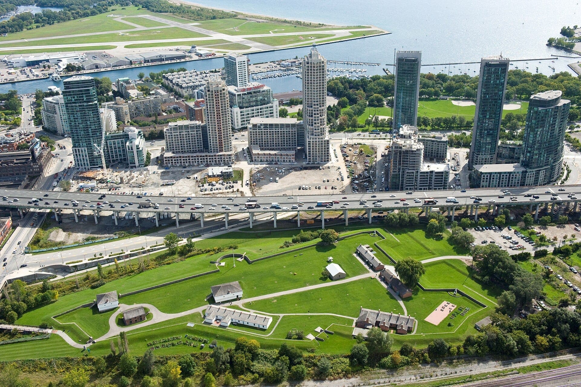 Vista aerea del Fort York National Historic Site con prati verdi, edifici storici e i grattacieli del centro di Toronto sullo sfondo in una giornata di sole.