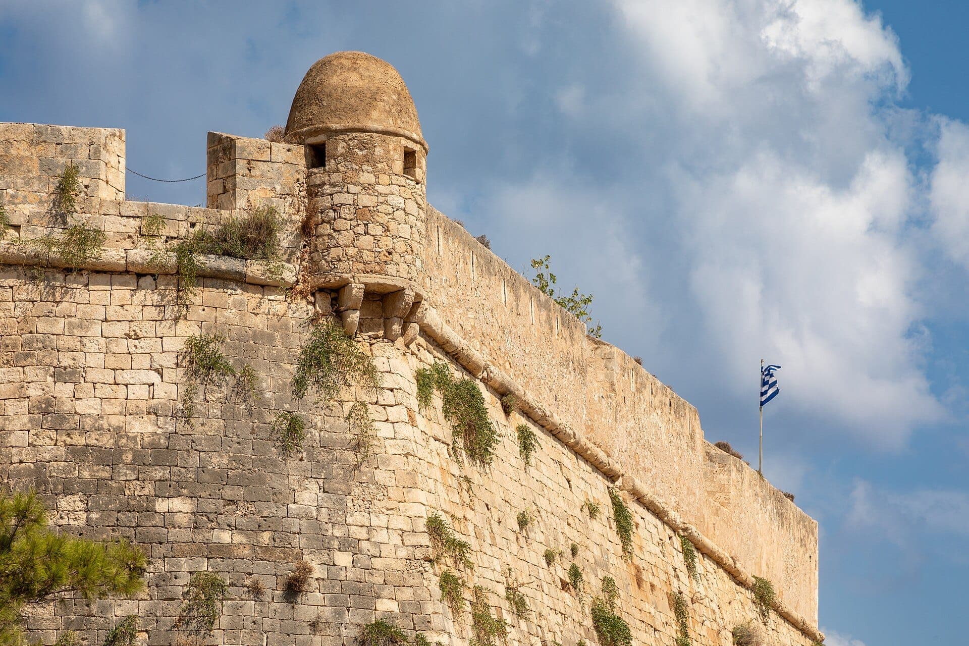 Steinmauern und ein Wachturm der Fortezza von Rethymno mit einer griechischen Flagge vor einem teilweise bewölkten Himmel.
