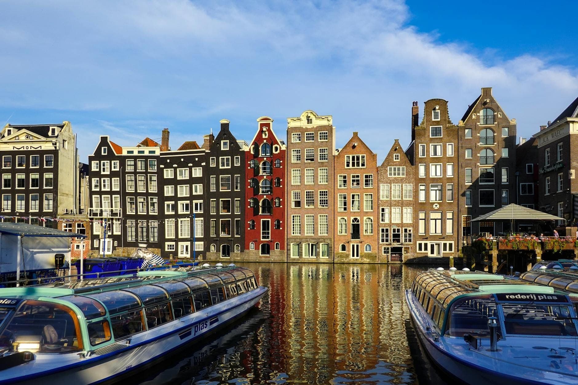 Klassische Amsterdamer Grachtenhäuser mit verzierten Giebeln, die sich im Wasser spiegeln, flankiert von Kanalbooten mit Glasdach unter einem strahlend blauen Himmel.